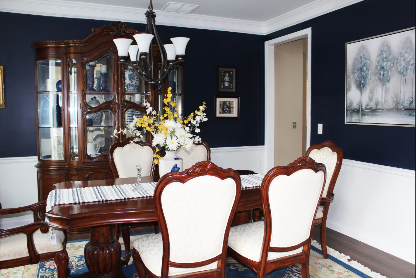 A formal dining room with a dark blue wall, a large wooden china cabinet, a wooden dining table with a striped table runner, six upholstered chairs with wooden frames, a floral centerpiece, a large painting of trees, and framed pictures on the wall.