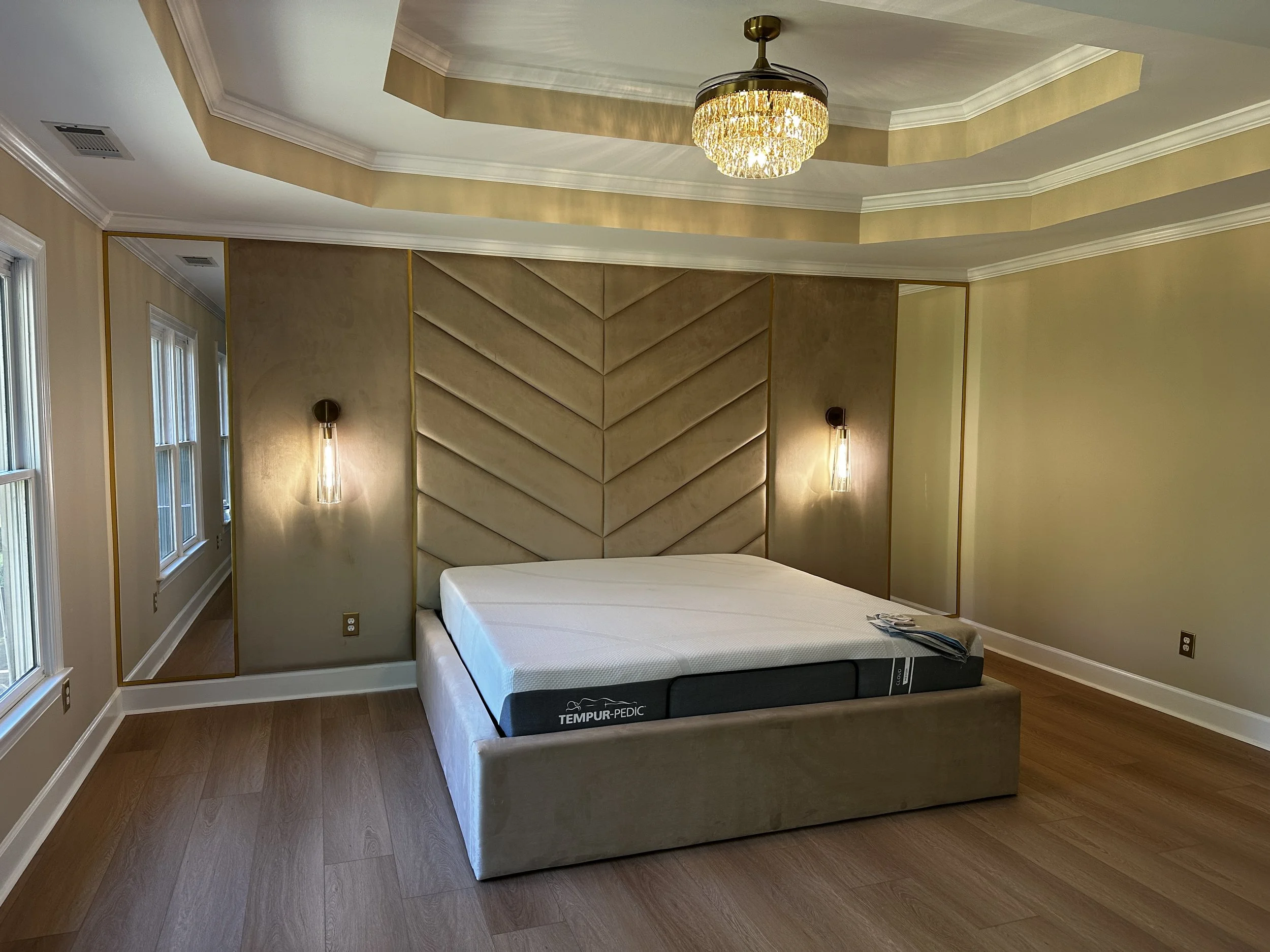 A bedroom with a bed, beige custom upholstered headboard, and a mattress with Tempur-Pedic branding, honey-colored wooden floor, windows on the left, and a gold and crystal chandelier hanging from the ceiling.