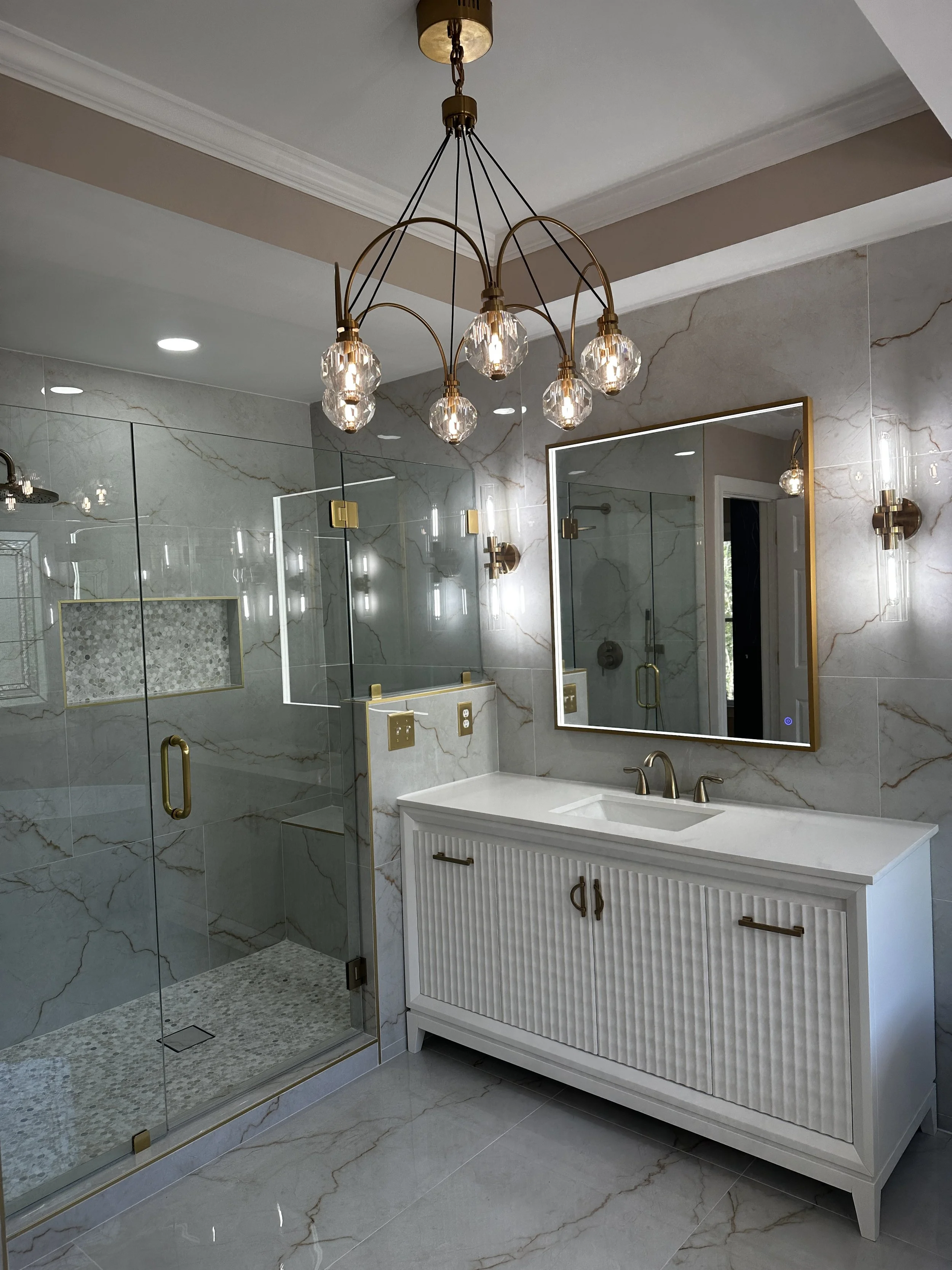 Modern bathroom with marble walls and flooring, glass shower with brass fixtures, white vanity with vertical slat detail, and wall-mounted lights
