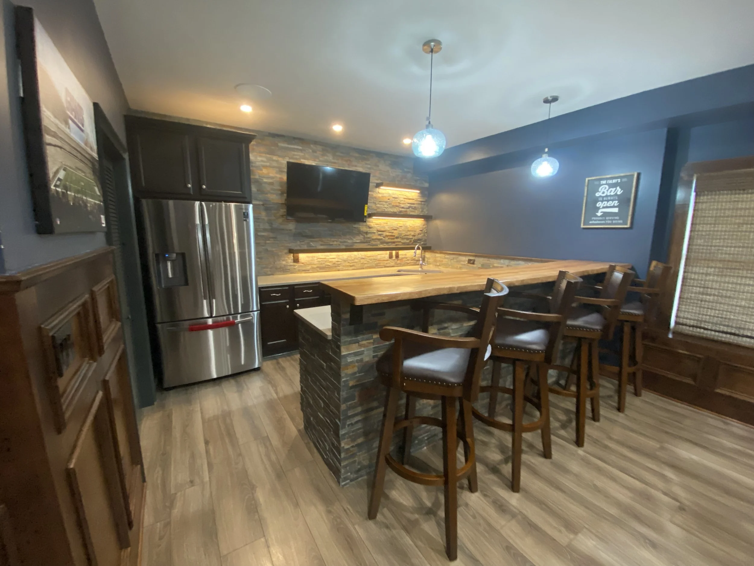 A basement kitchen with dark cabinets, a stainless steel refrigerator, a stone accent wall with a mounted TV, hanging pendant lights, a wooden countertop island with bar stools, and a blue wall with a framed sign.