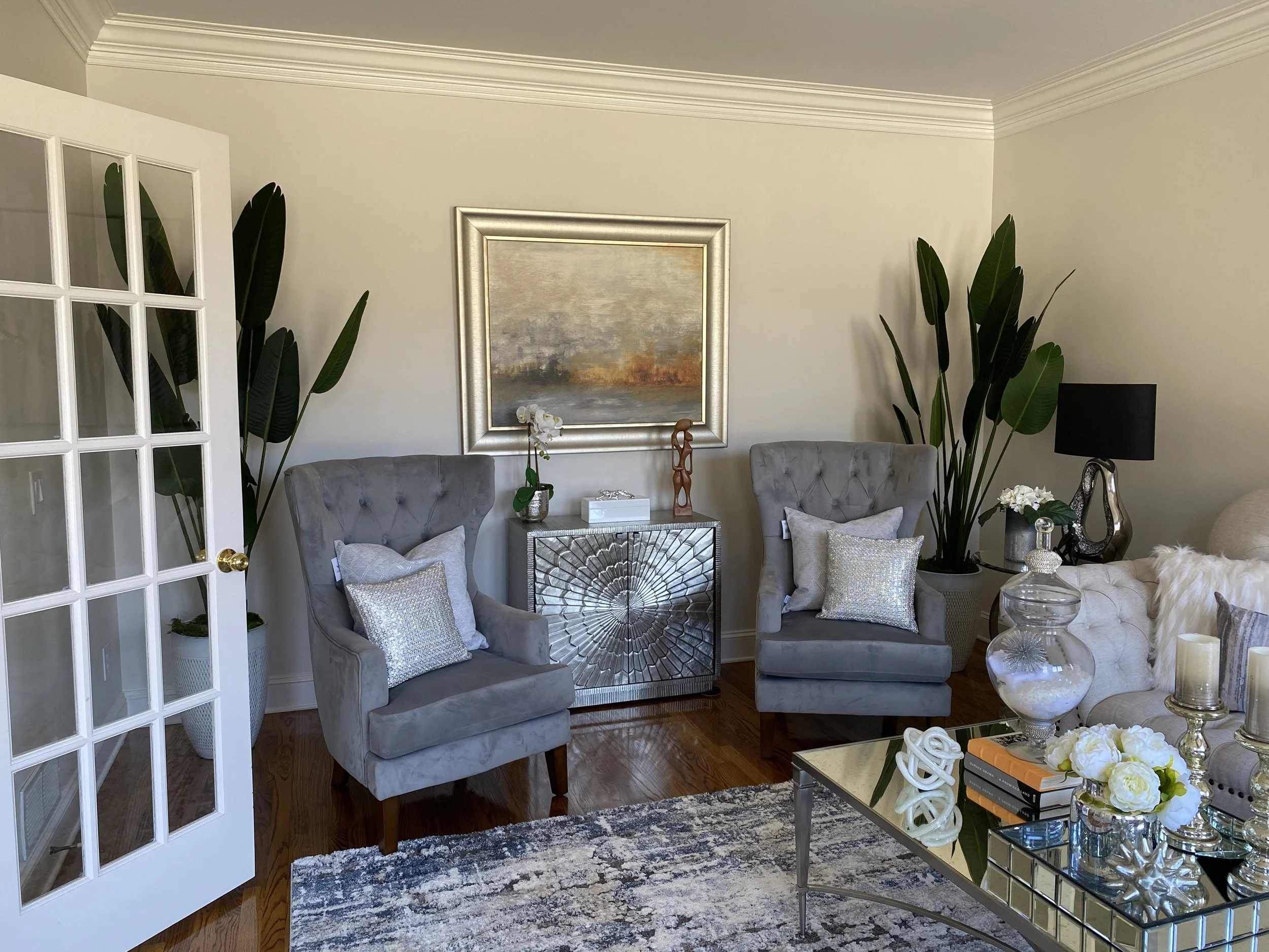 A cozy living room corner with two gray upholstered armchairs with white pillows, a mirrored side table with decorative objects, a large framed landscape painting, tall green plants, and a glass coffee table with flowers and books.