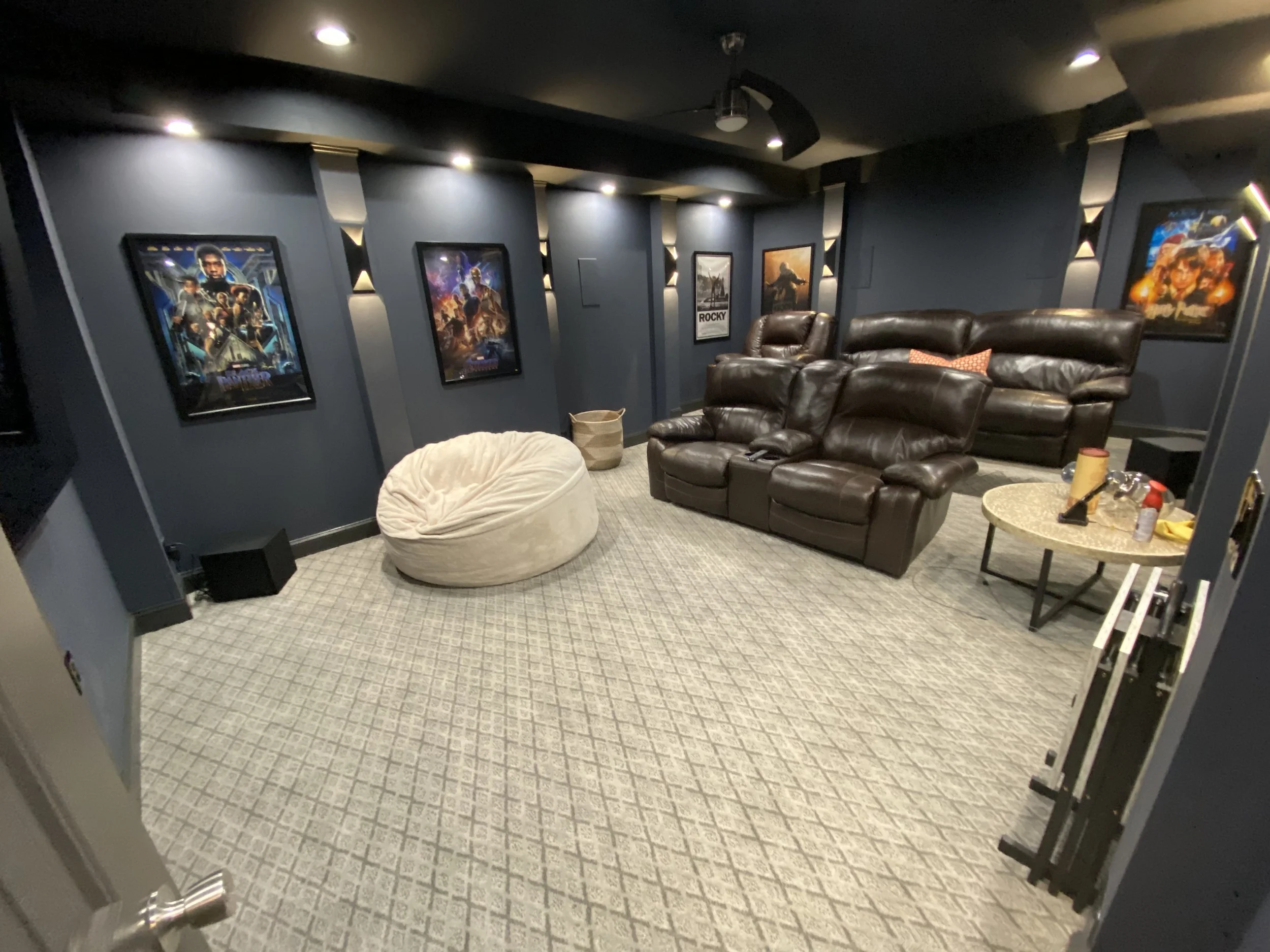 A cozy home theater room with dark blue walls, decorated with framed movie posters, including Marvel, Guardians of the Galaxy, Rocky, and Star Wars. The room has leather recliner seating, a bean bag chair, a coffee table with snacks, and small decora