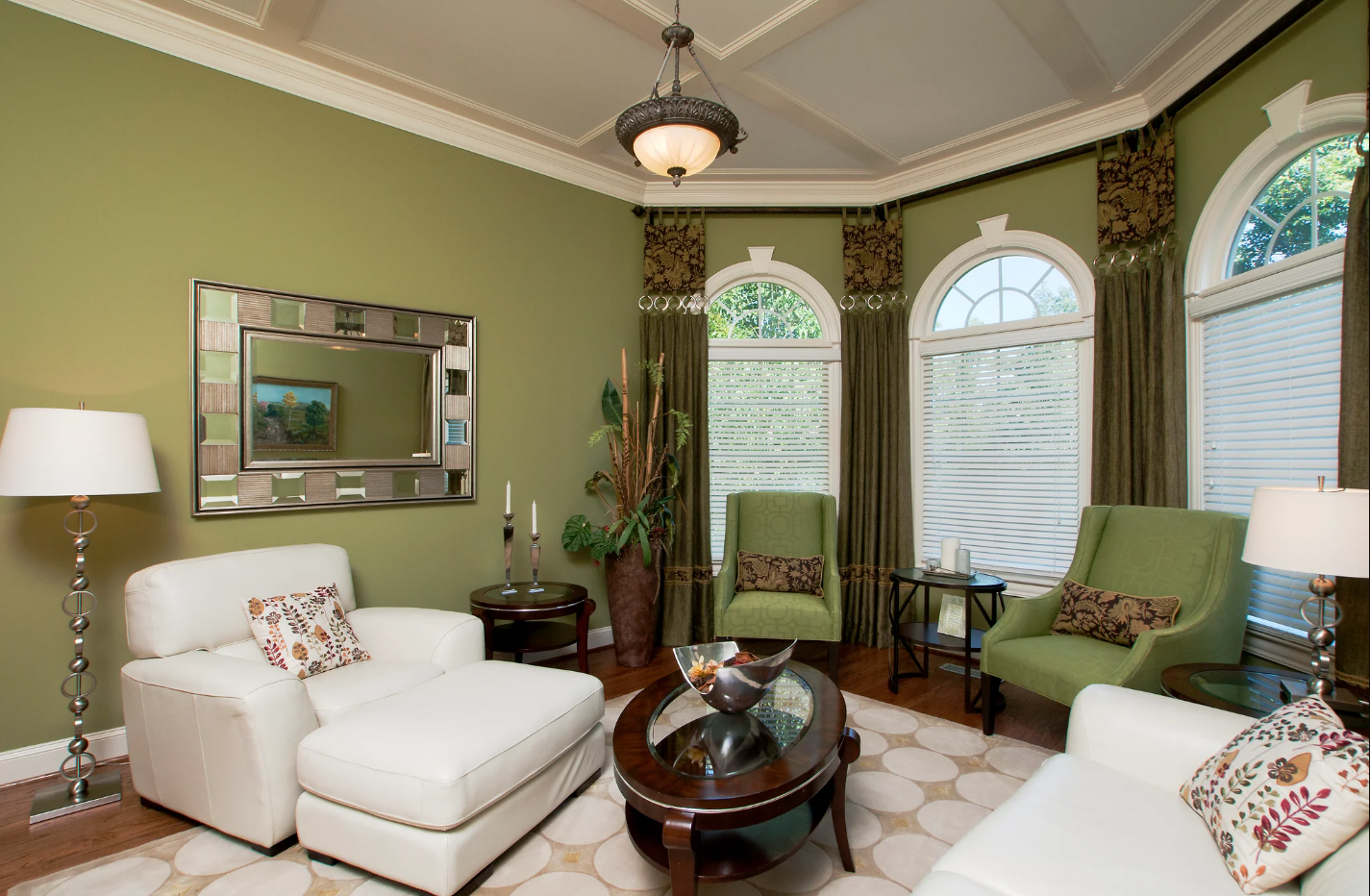 Living room with green walls, large arched windows with blinds and curtains, two white sofas, two green armchairs, table with candles, decorative bowl, lamp, mirror, and indoor plant.