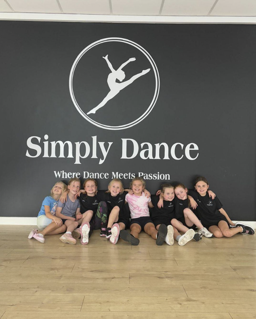 Group of young girls sitting on the floor in front of a wall that says "Simply Dance" with a logo of a dancer, and the tagline "Where Dance Meets Passion."