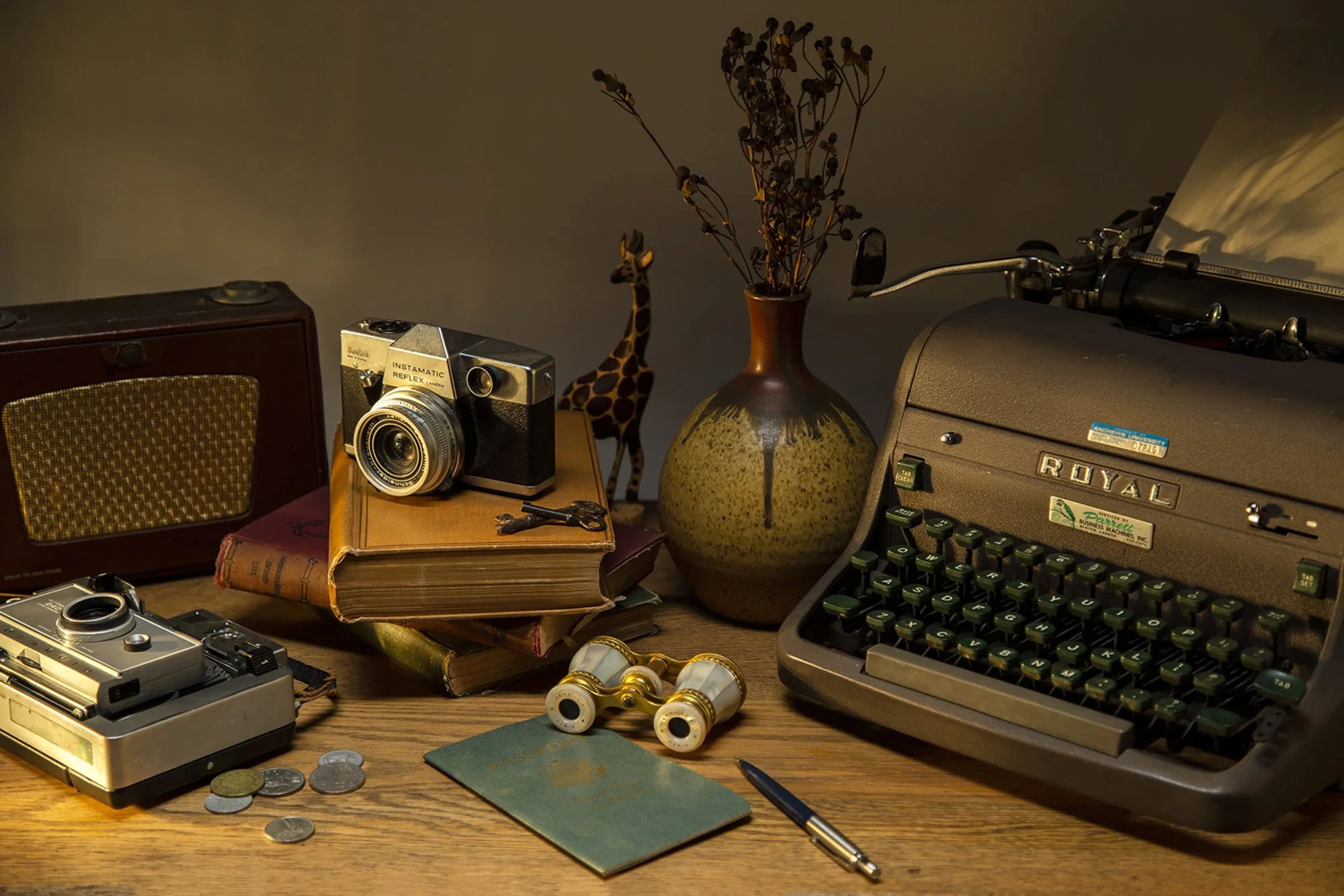 A vintage desk with old-fashioned camera equipment, coins, a small notebook, and a fountain pen. The scene includes a ceramic giraffe figurine, a vase with dried flowers, an old typewriter, and a compact camera. The lighting is warm and subdued, crea