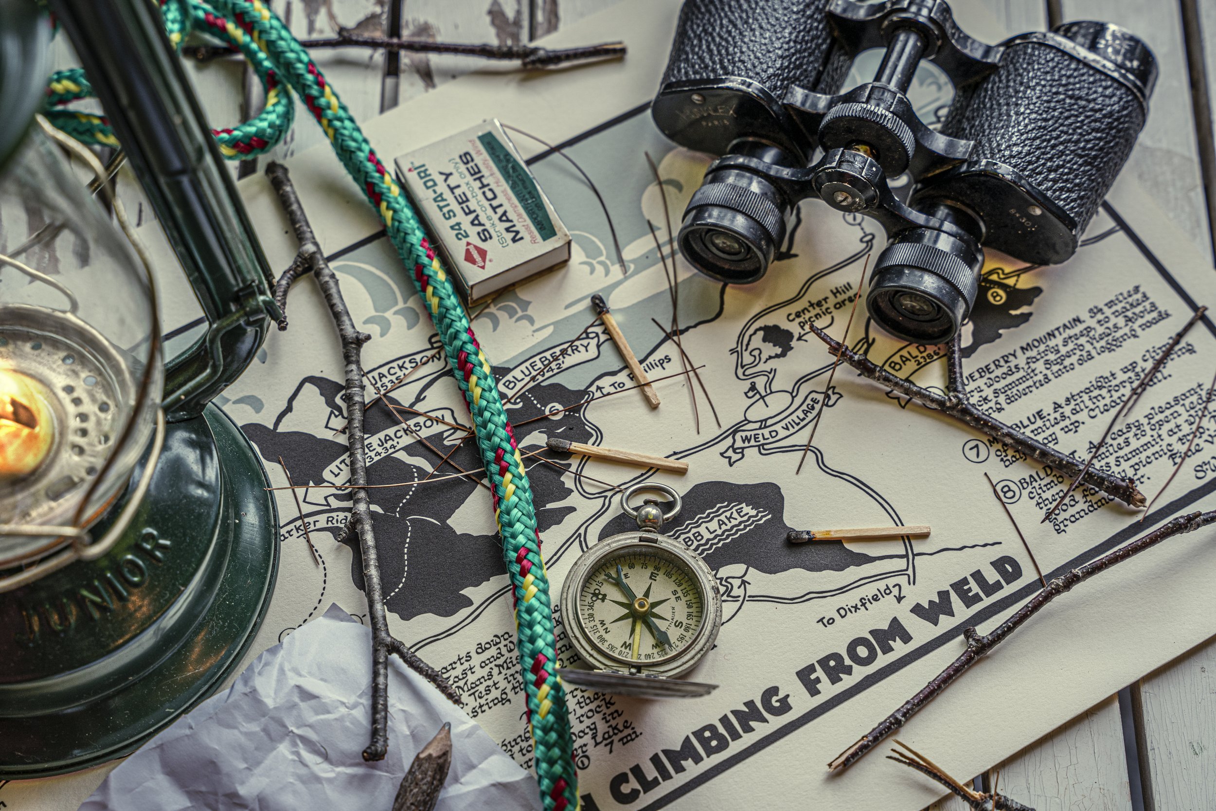 Close-up photo of binoculars, a compass, a matchbox, a colorful rope, a lantern, and a map with marked trails and text, suggesting an outdoor adventure or exploration scene.