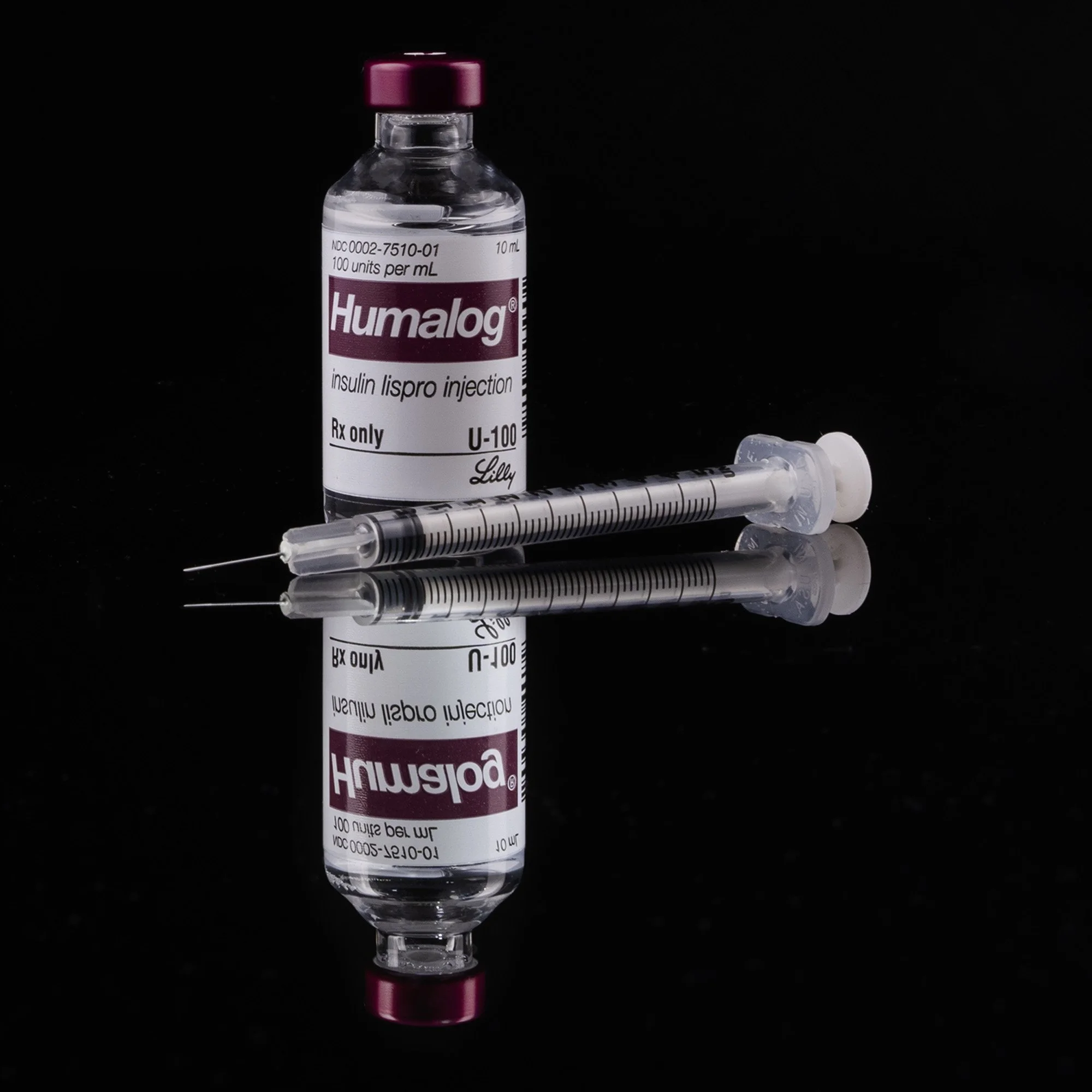 A vial of Humalog insulin with a syringe placed below on a reflective surface against a black background.