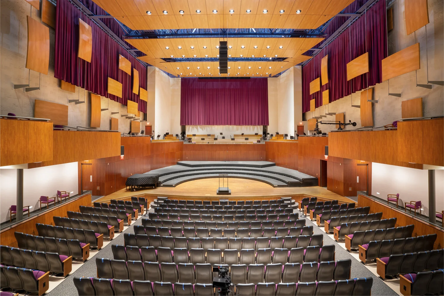 Empty auditorium with tiered seating, a stage with a podium, and large red curtains.