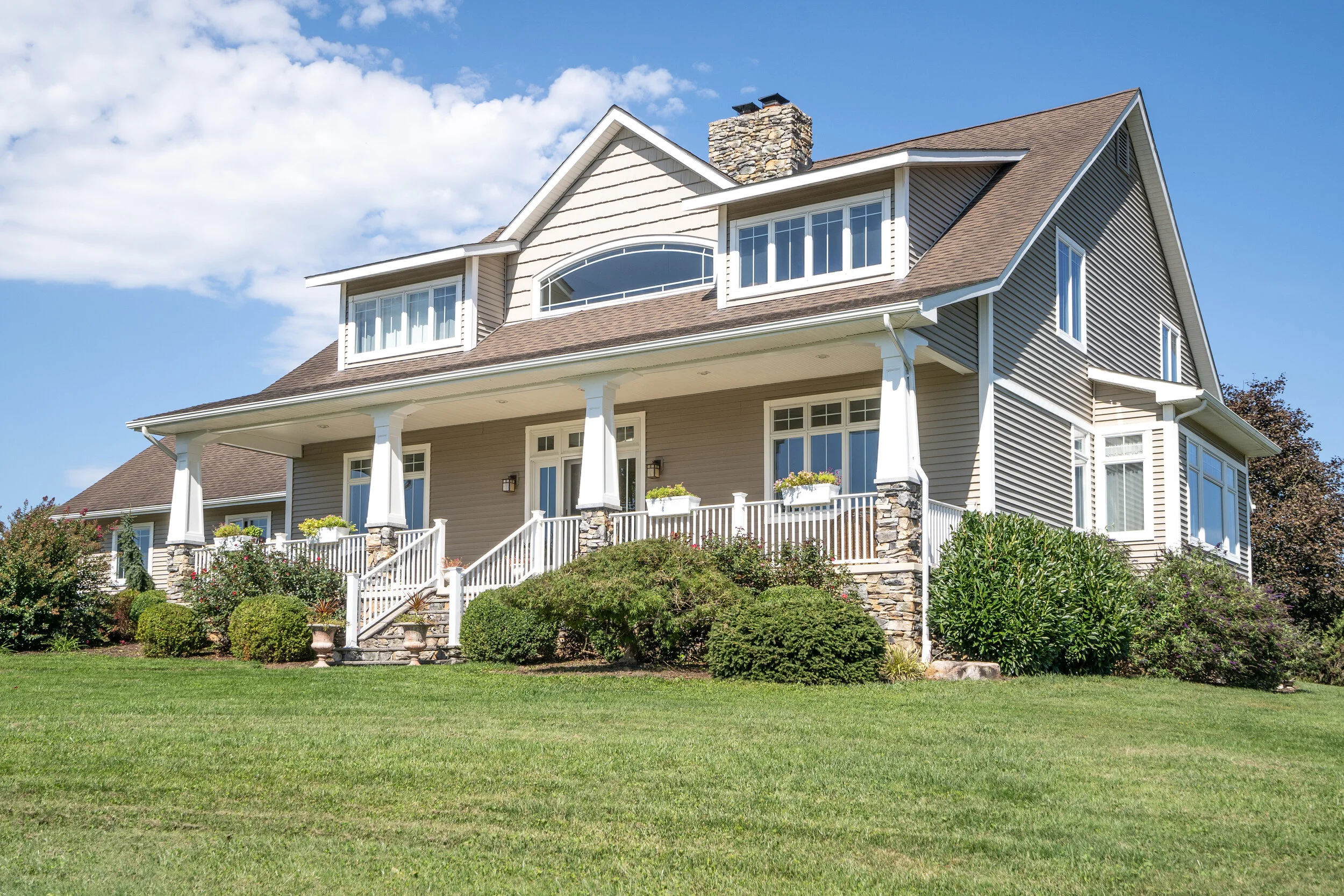 A large two-story house with beige siding, white trim, and multiple windows, surrounded by a well-manicured lawn and landscaped garden with bushes and flowering plants, under a blue sky with some clouds.
