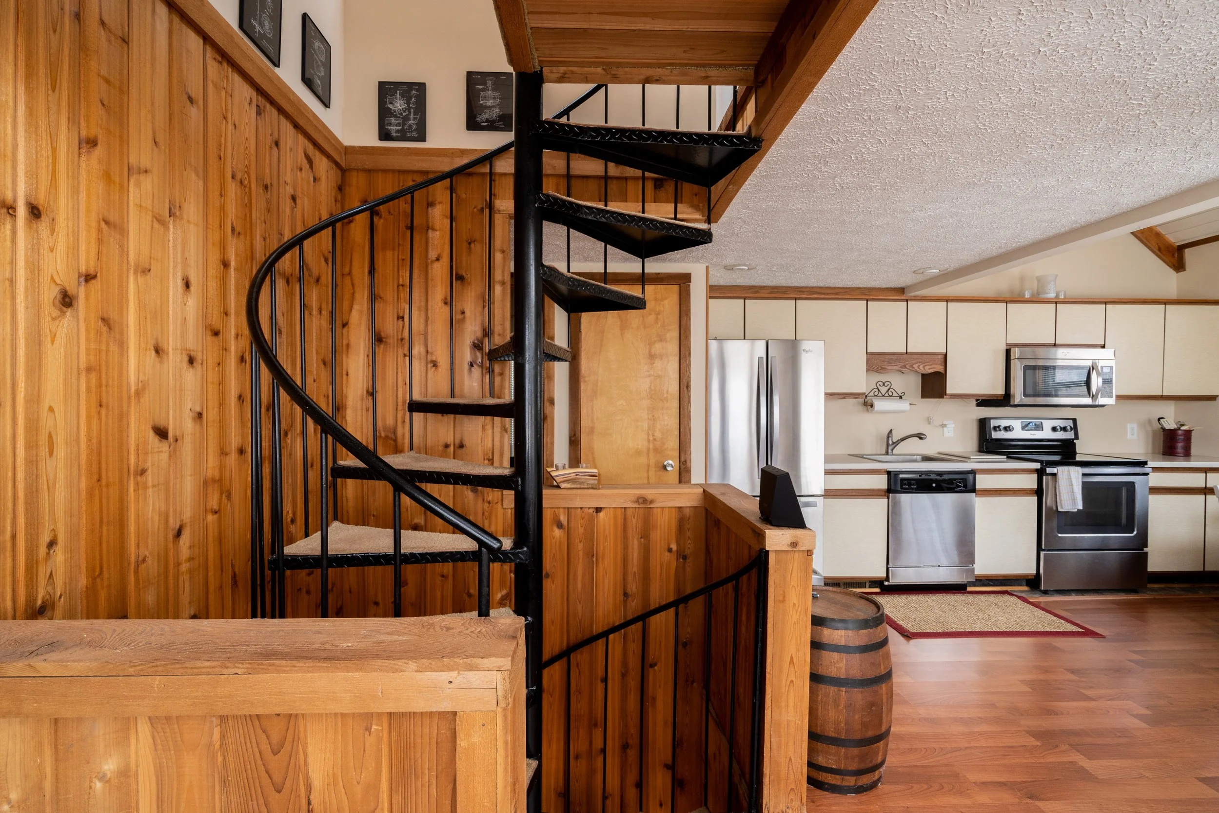 Interior of a kitchen with cream-colored cabinets, stainless steel appliances, and a spiral staircase with black metal frame and wooden steps, adjacent to wood-paneled walls and ceiling.