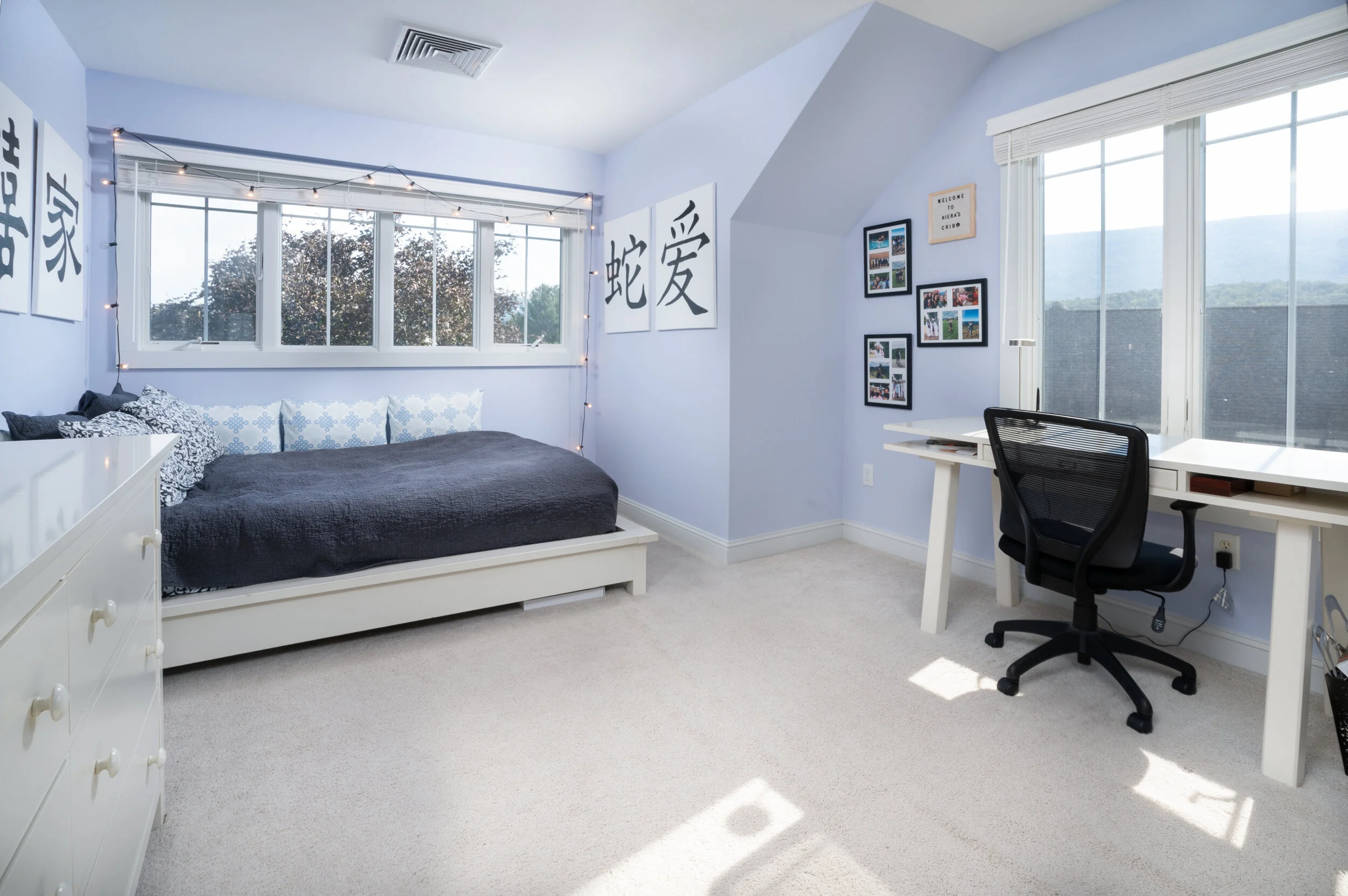 A bedroom with light blue walls, featuring a bed with black and white pillows, large windows with blinds, and framed wall art including Asian characters and photographs, with a white desk and black chair near a window.