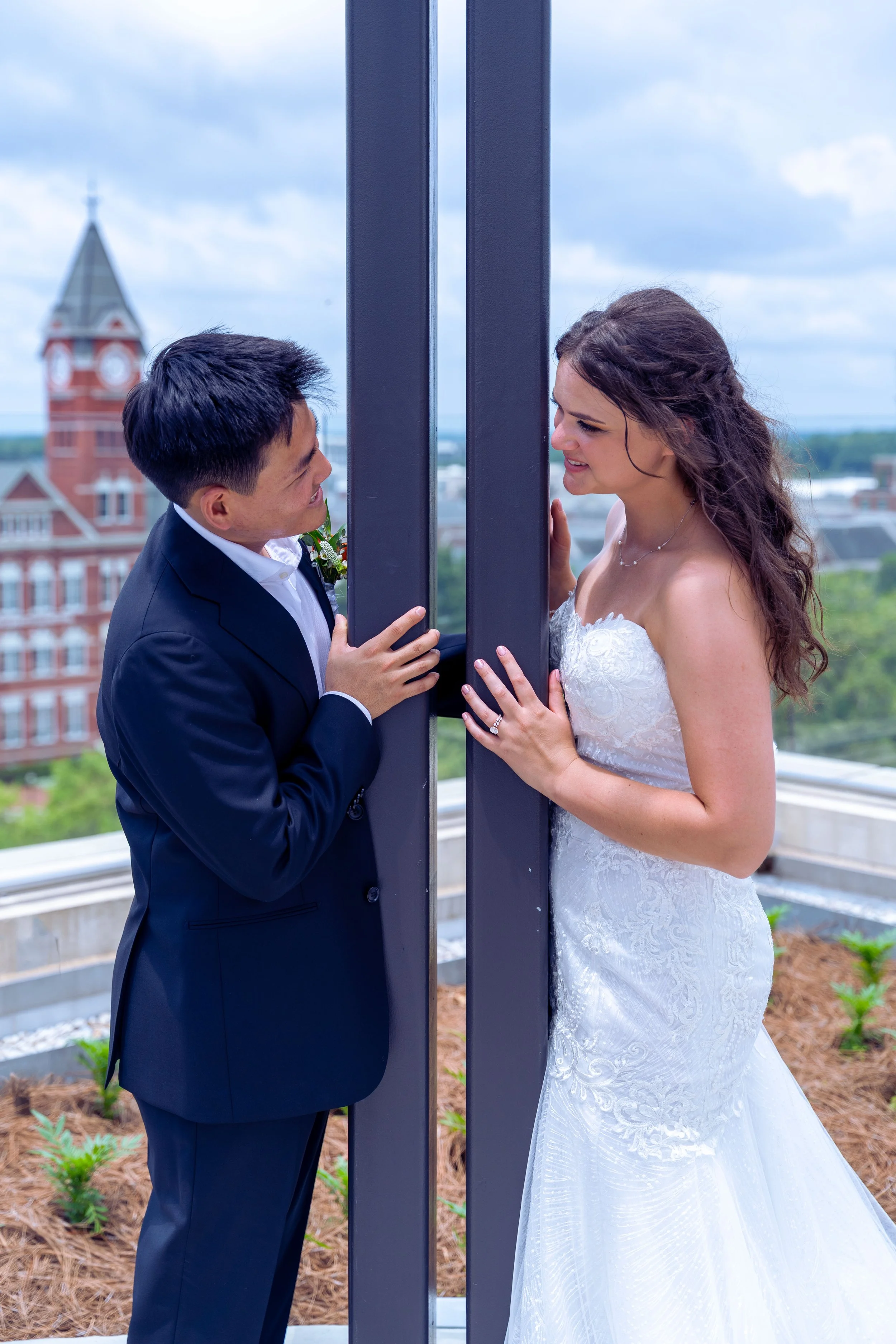 A couple, dressed in wedding attire, stands on either side of a vertical black beam, touching it and gazing at each other, on an outdoor balcony with a cityscape background including a red brick building with a clock tower.