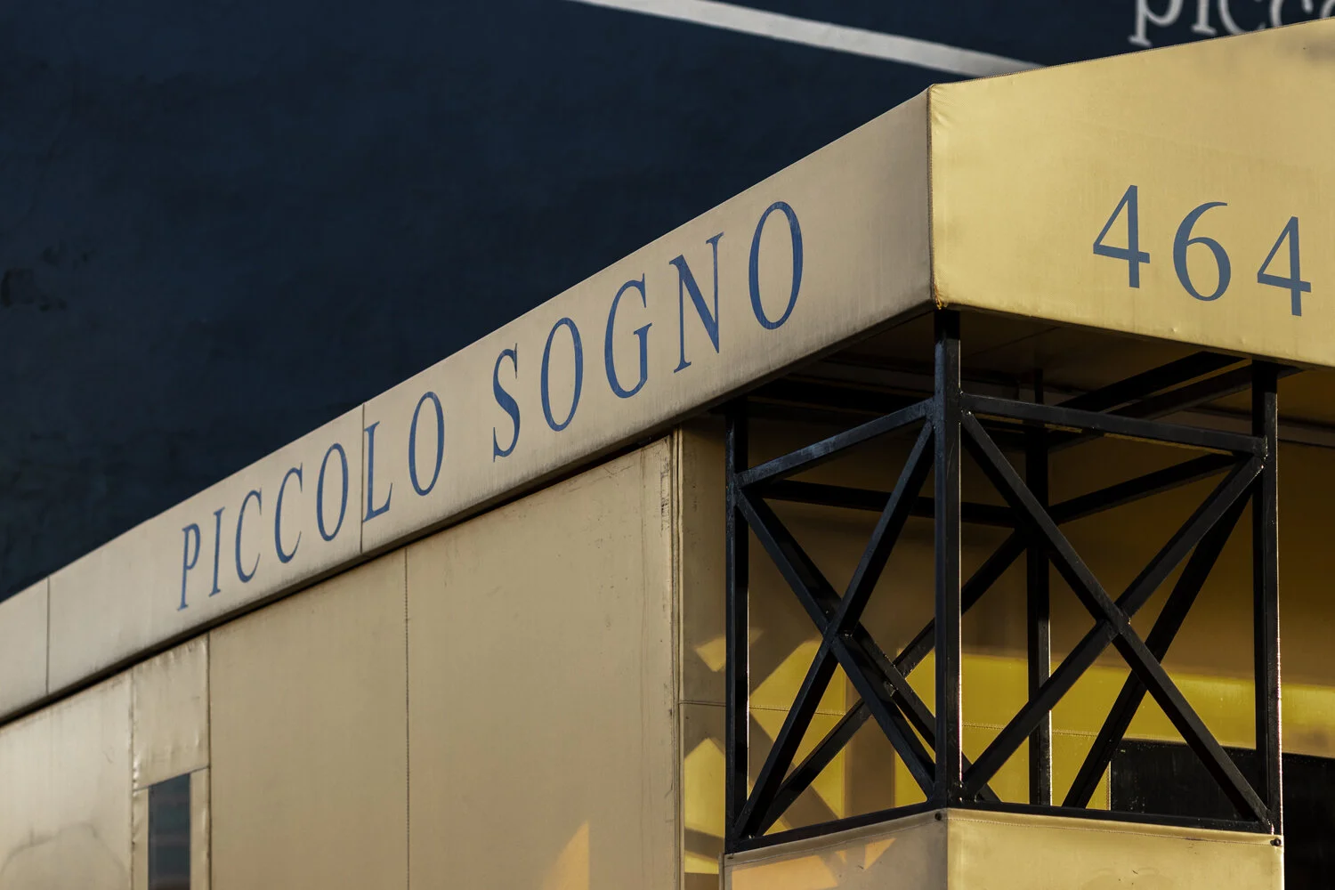 Close-up of a building corner with the name 'PICCOLO SOGNO' and the number 464, showing a beige facade, black metal balcony, and dark sky in the background.