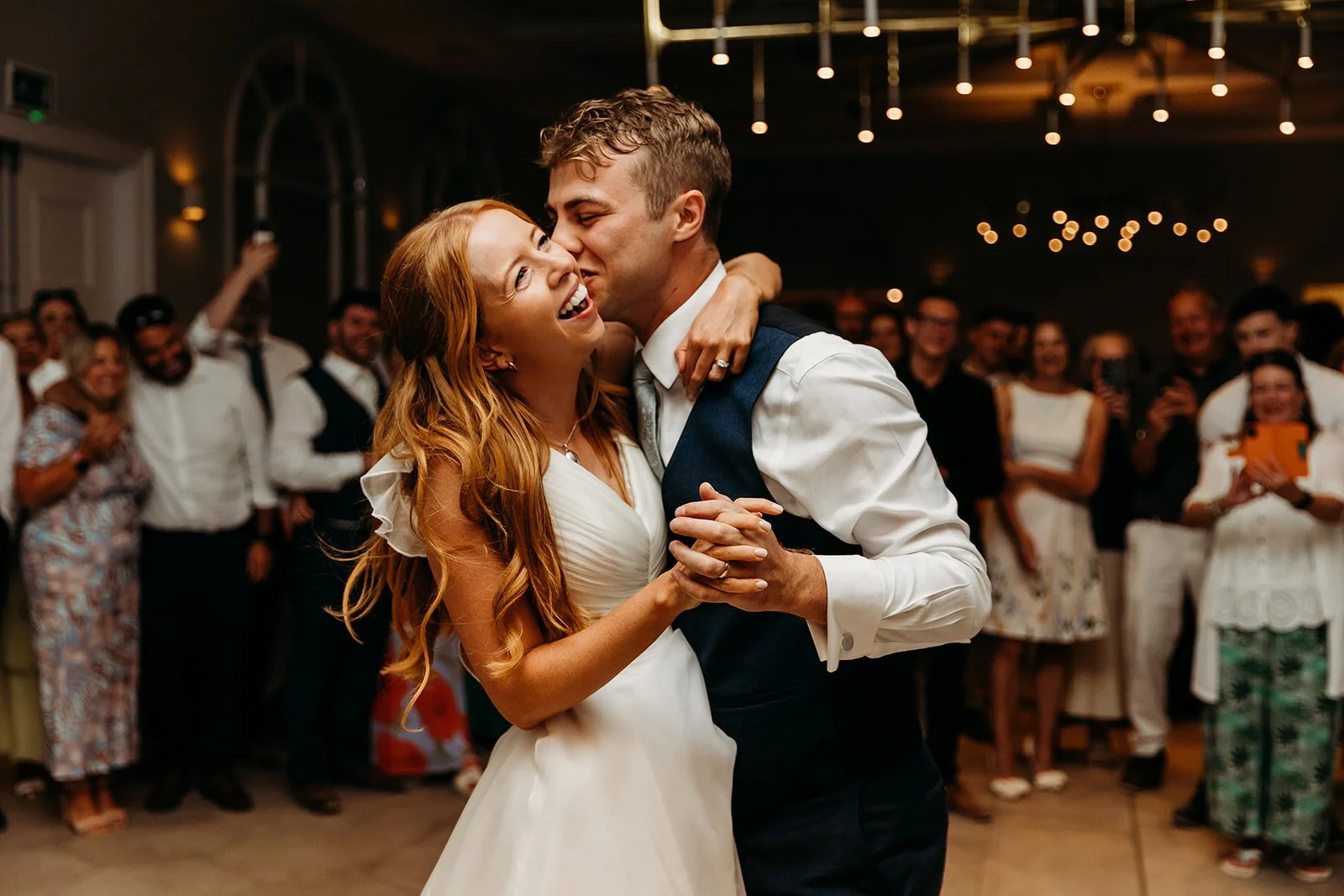 A bride and groom are dancing closely, smiling, and sharing a joyful moment during their wedding reception, with guests clapping and taking photos in the background.