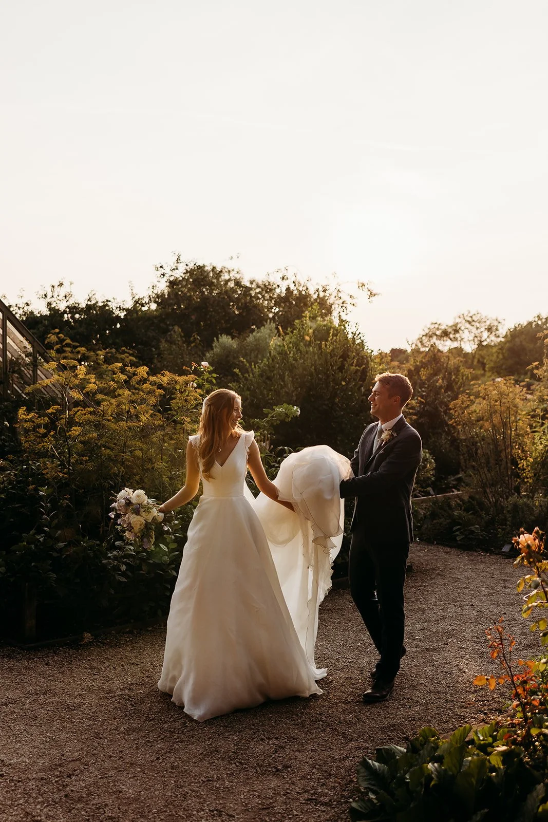 Bride and groom holding wedding dress and bouquet in a garden during sunset at deer park country house
