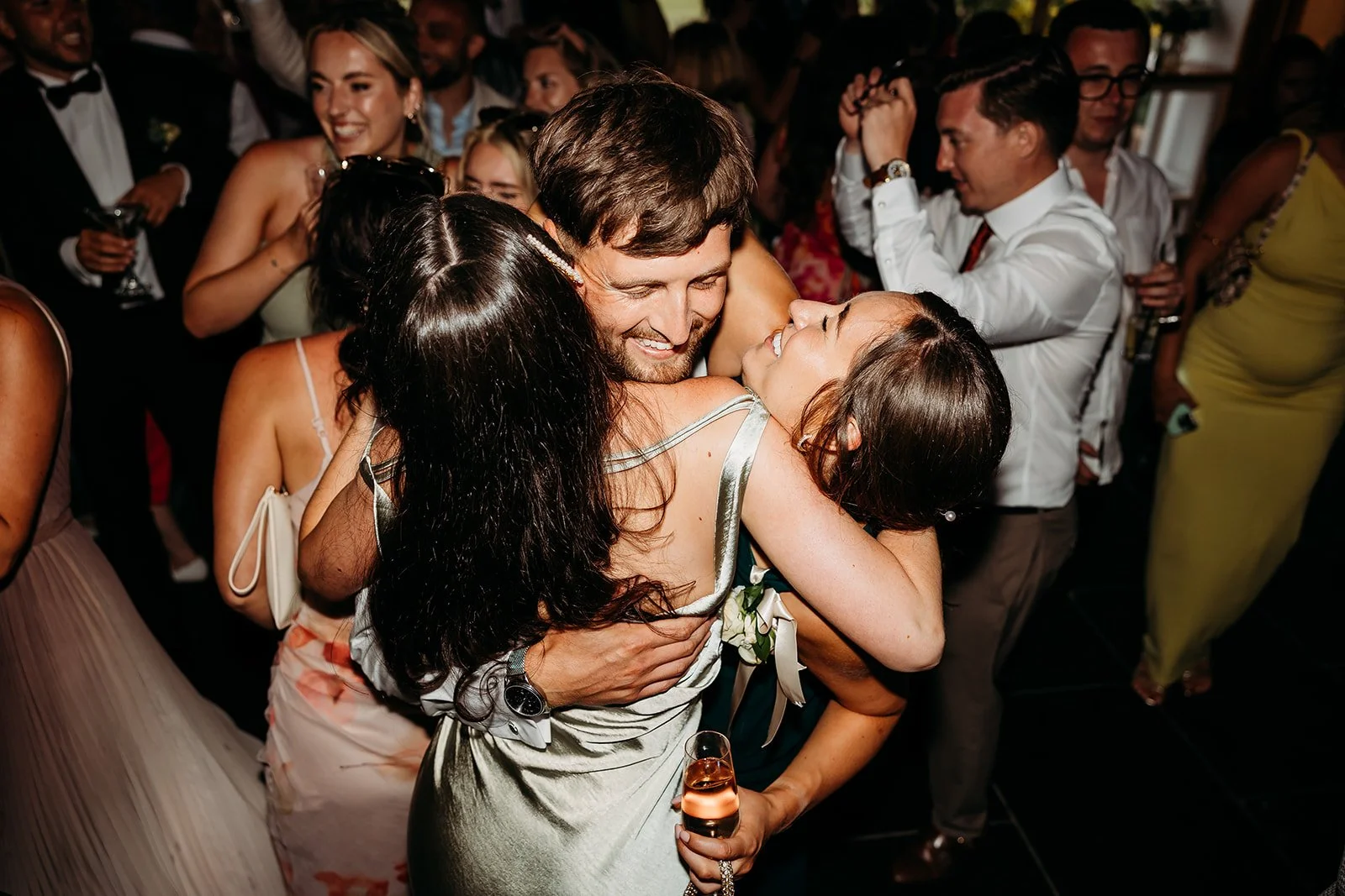 People dancing and hugging at a celebration or party, with some holding drinks, in a lively and joyful atmosphere.