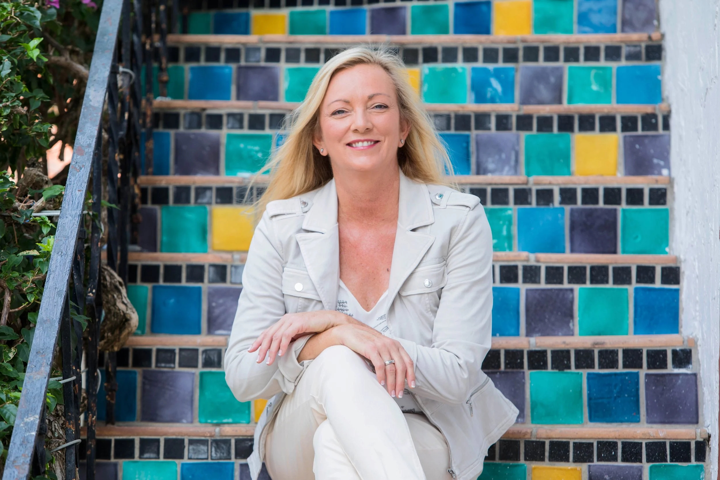 A woman with blonde hair in a light-colored jacket and pants sitting on colorful tiled stairs, smiling at the camera.