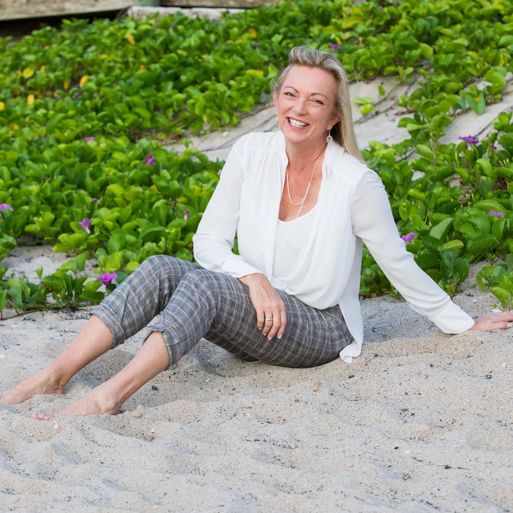 A smiling woman with blonde hair, dressed in a white shirt and plaid pants, sitting barefoot on the sand at the beach near green plants with purple flowers.