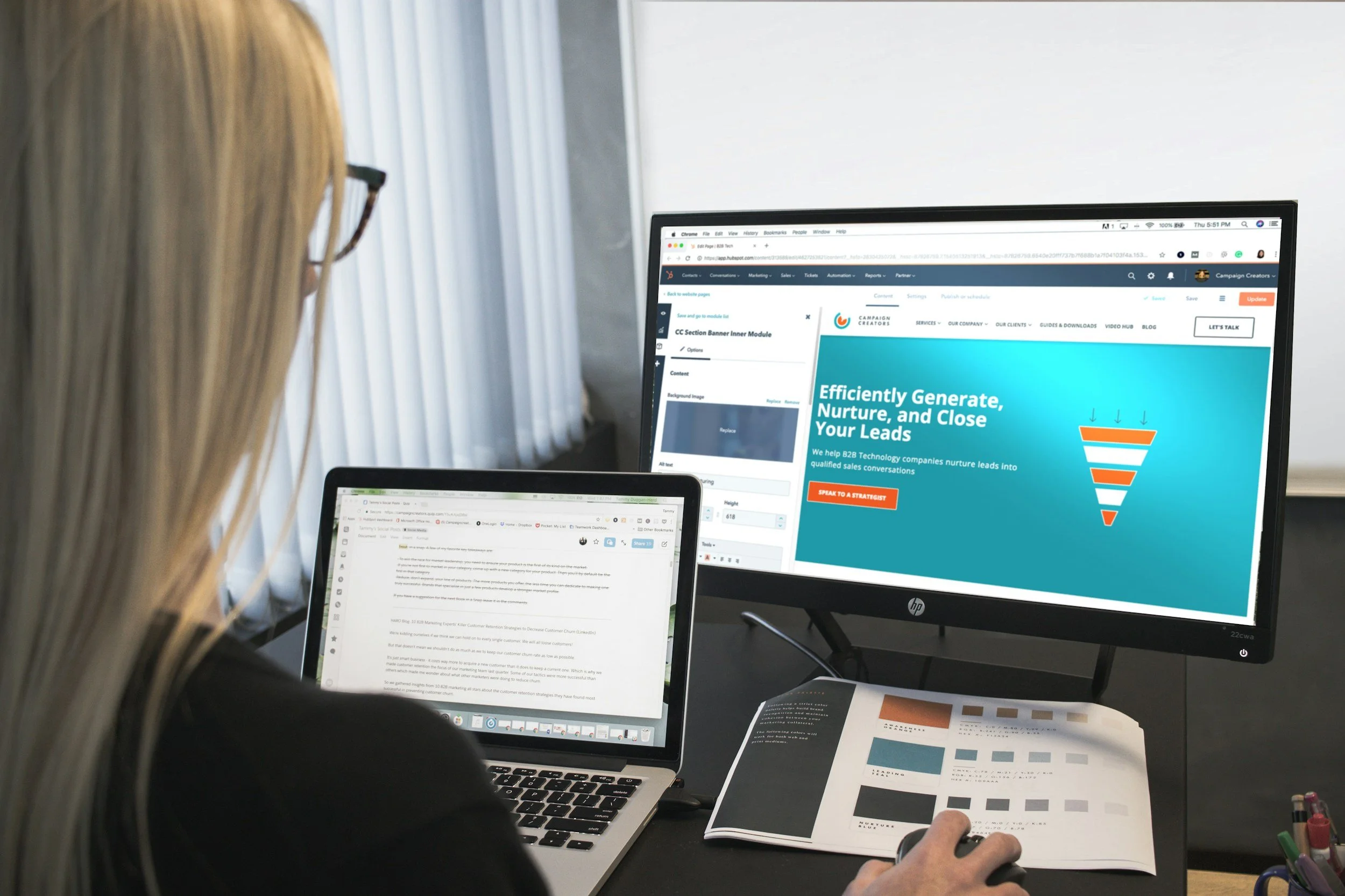A woman with long blonde hair and glasses working at a desk with a laptop, an external monitor, a color swatch booklet, and a pen holder.