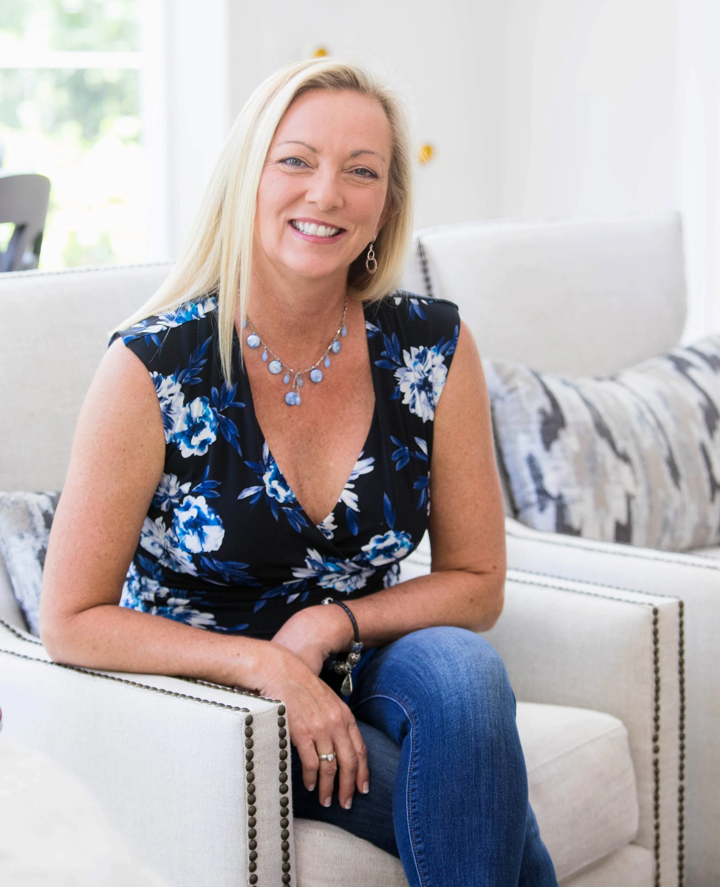A woman with blonde hair smiling, sitting on a cream-colored sofa with patterned pillows, wearing a navy floral sleeveless top and blue jeans, in a bright room with a window in the background.