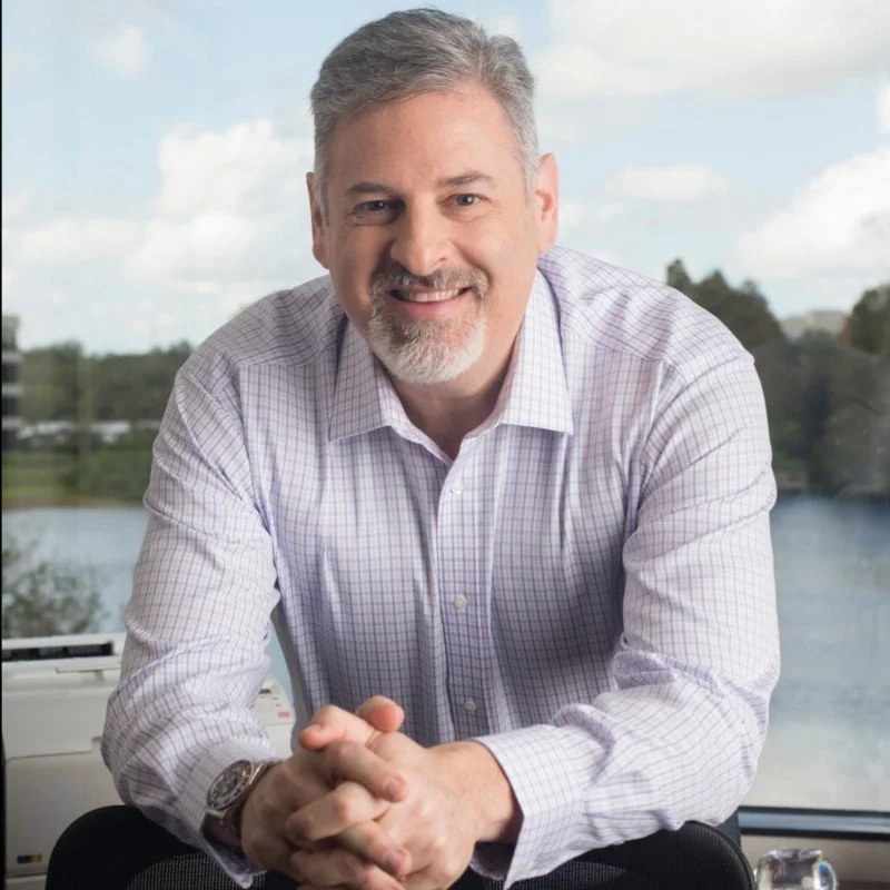 A smiling middle-aged man with gray hair and a beard, wearing a light-colored checkered dress shirt, sitting indoors with a window view of a river, trees, and a partly cloudy sky in the background.