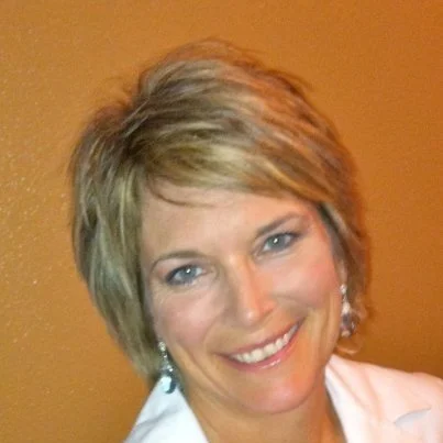 Close-up of a smiling woman with short, layered blonde hair, wearing earrings and a white top, against a brown wall.