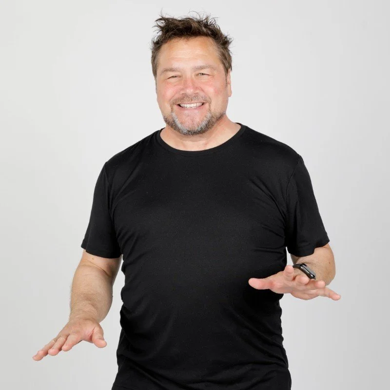 Man with short, curly hair and a beard, wearing a black t-shirt, smiling and gesturing with his hands, standing against a white background.