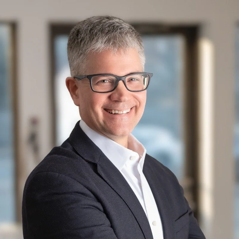 A smiling man with short gray hair, glasses, wearing a white shirt and black blazer, standing indoors with blurred windows in the background.