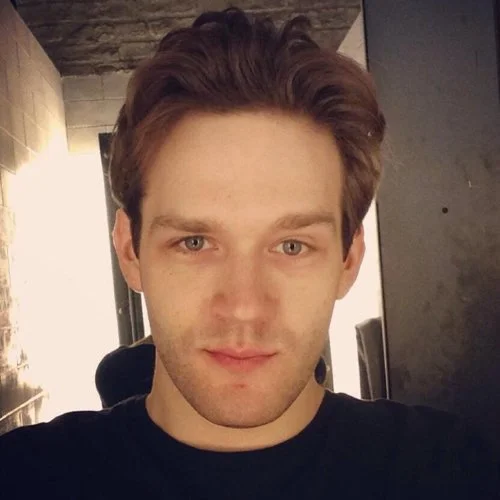 Close-up of a young man with brown hair, wearing a black shirt, in an indoor setting.
