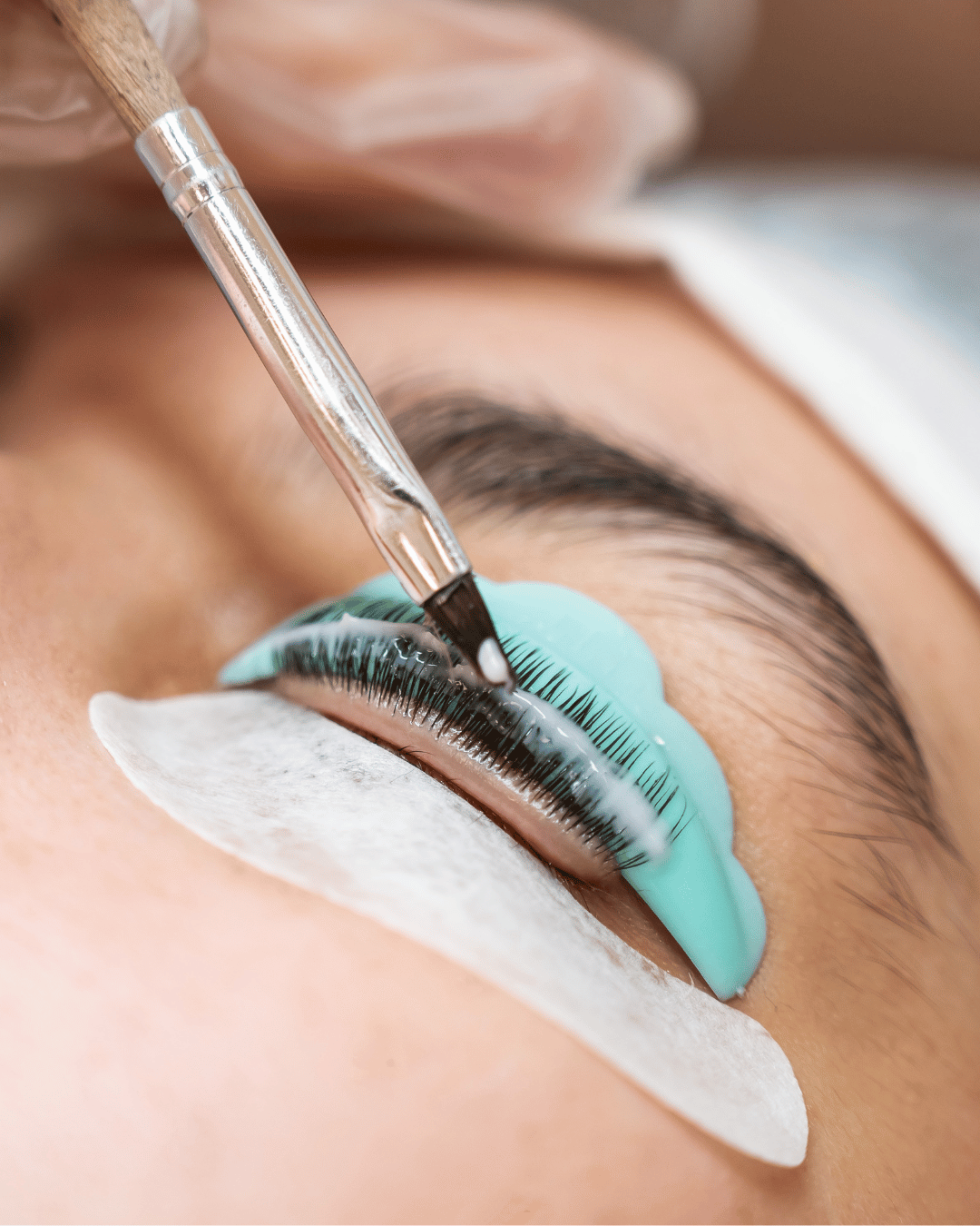Close-up of a person receiving eyelash extension application, with a technician using a fine brush and blue adhesive on the eyelid, eyelash pads under the eye for protection.