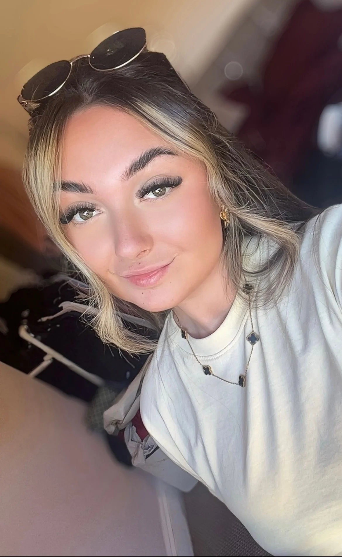 A woman with long dark hair tied in a ponytail, wearing a black shirt, is sitting in a white chair with gold accents, looking down and slightly to her left, beside a girl lying on a bed, with a curtain partially visible, in a room with white walls and a mirror in the background.