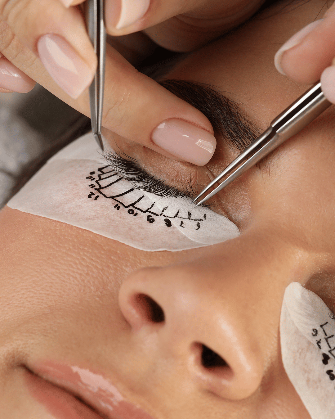 A person is performing eyelash extension application, with a technician using tweezers to apply individual lashes to the client's upper eyelid, which is covered with a protective pad.
