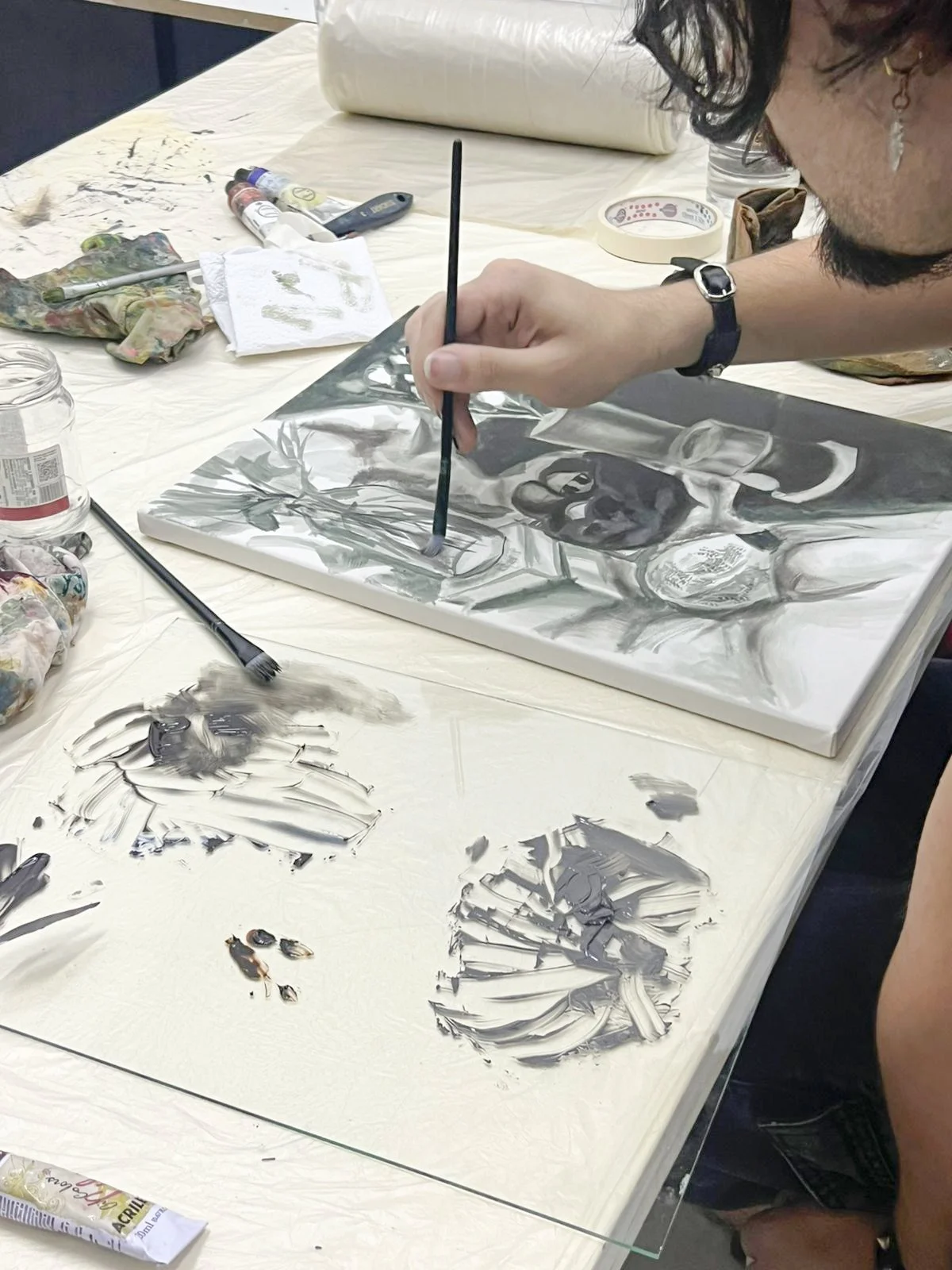 Artista aluno pintando uma tela em uma mesa de trabalho, com vários tubos de tinta, pincéis, pano e outros materiais de pintura ao redor.