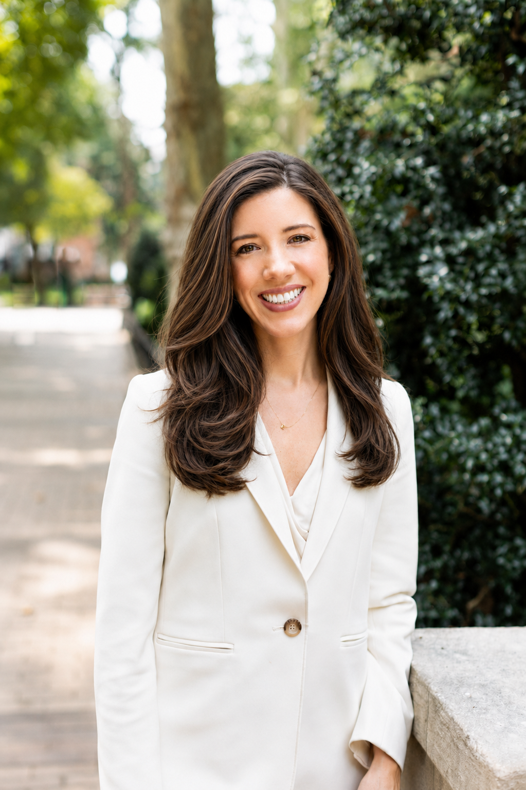 Smiling woman with long brown hair in a navy dress standing outdoors in a green leafy setting.