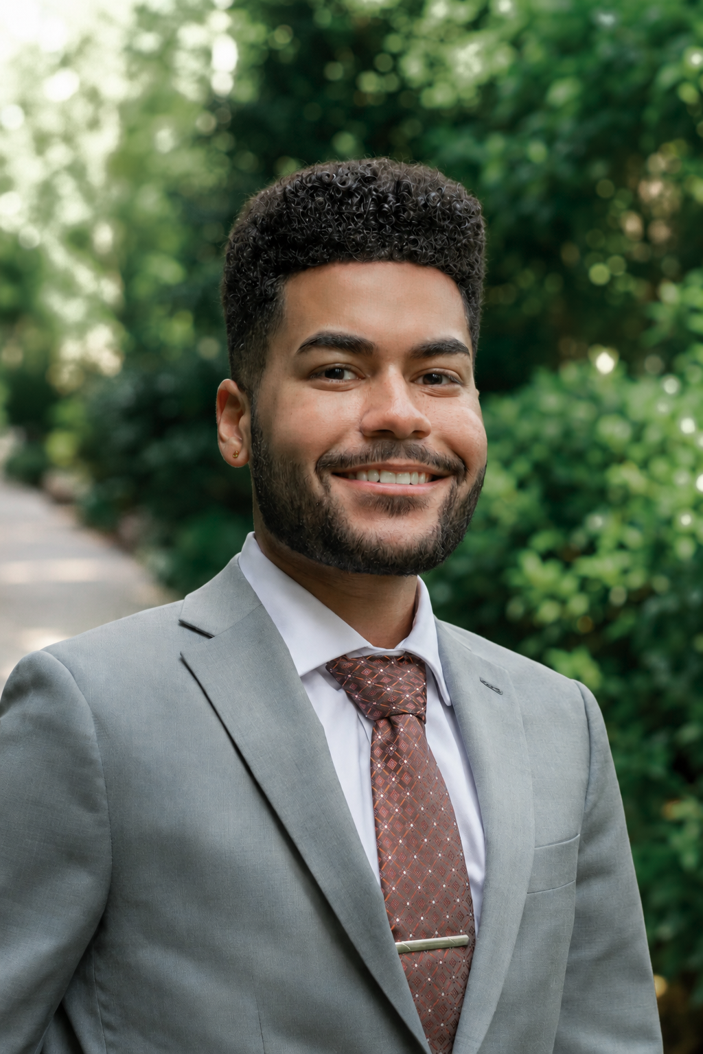A confident person in a suit and floral tie smiling. Background features greenery and blurred outdoor setting.