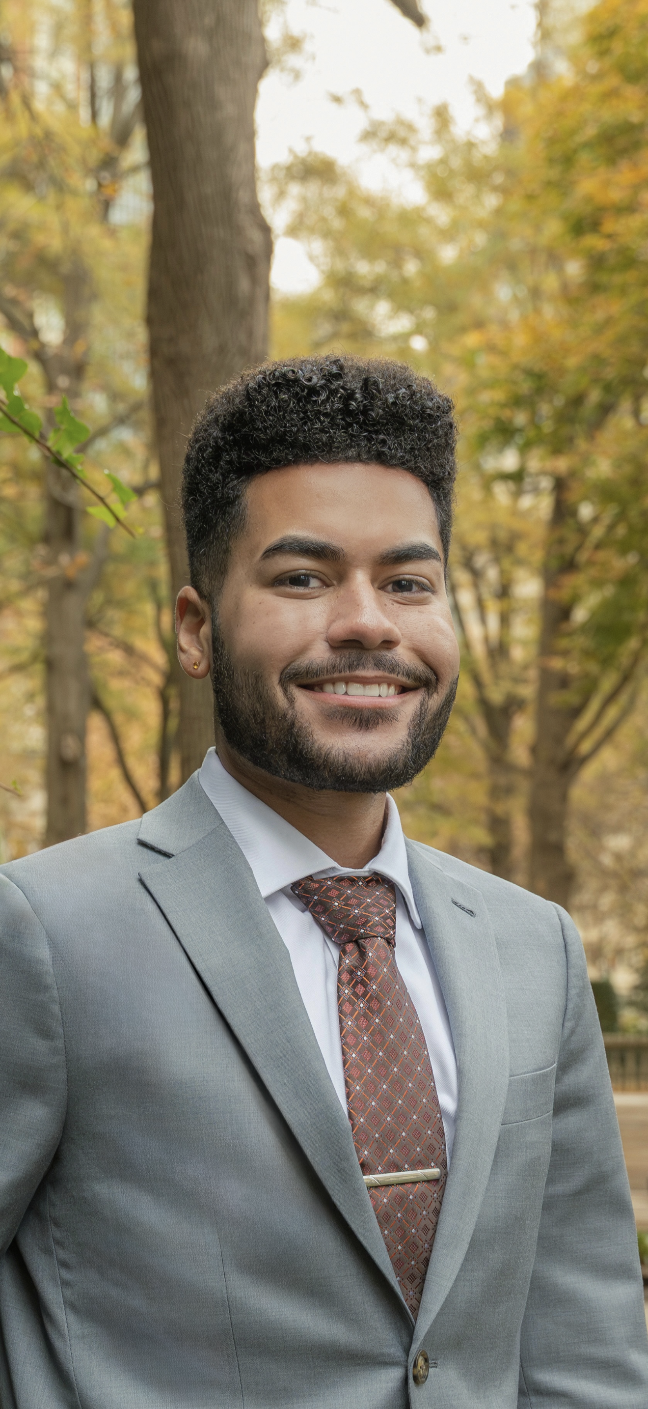 A confident person in a suit and floral tie smiling with arms crossed. Background features greenery and blurred outdoor setting.