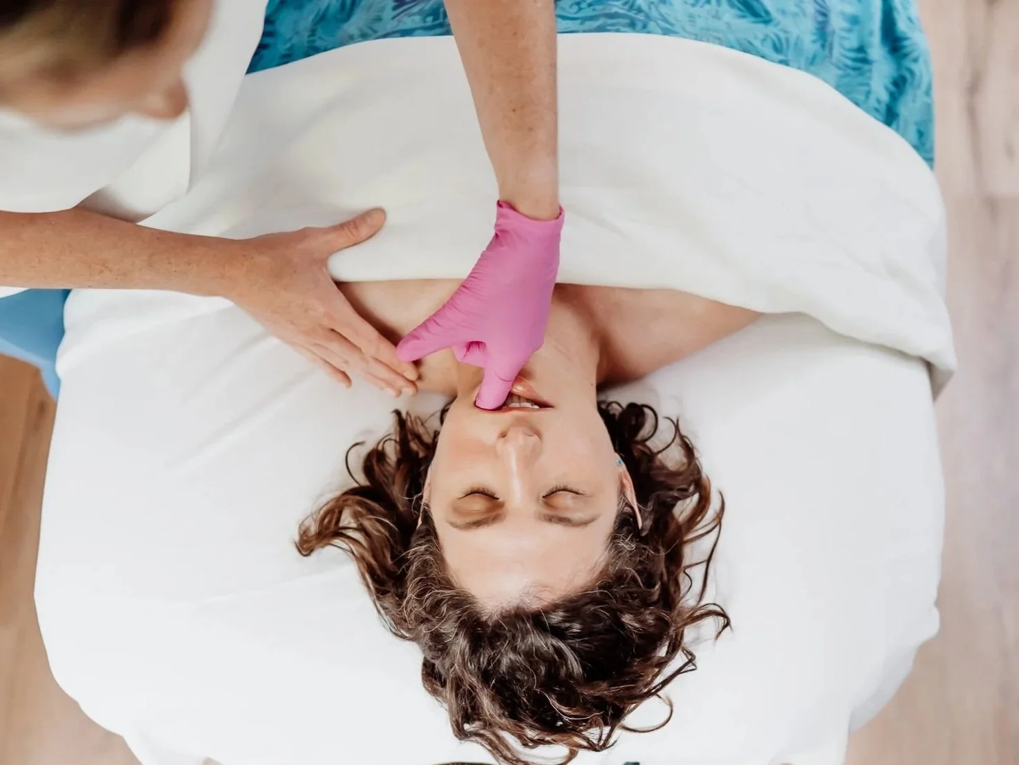 Client receiving intra-oral TMJ massage on clean, bright white sheets while the massage therapist uses a medical glove inside the mouth