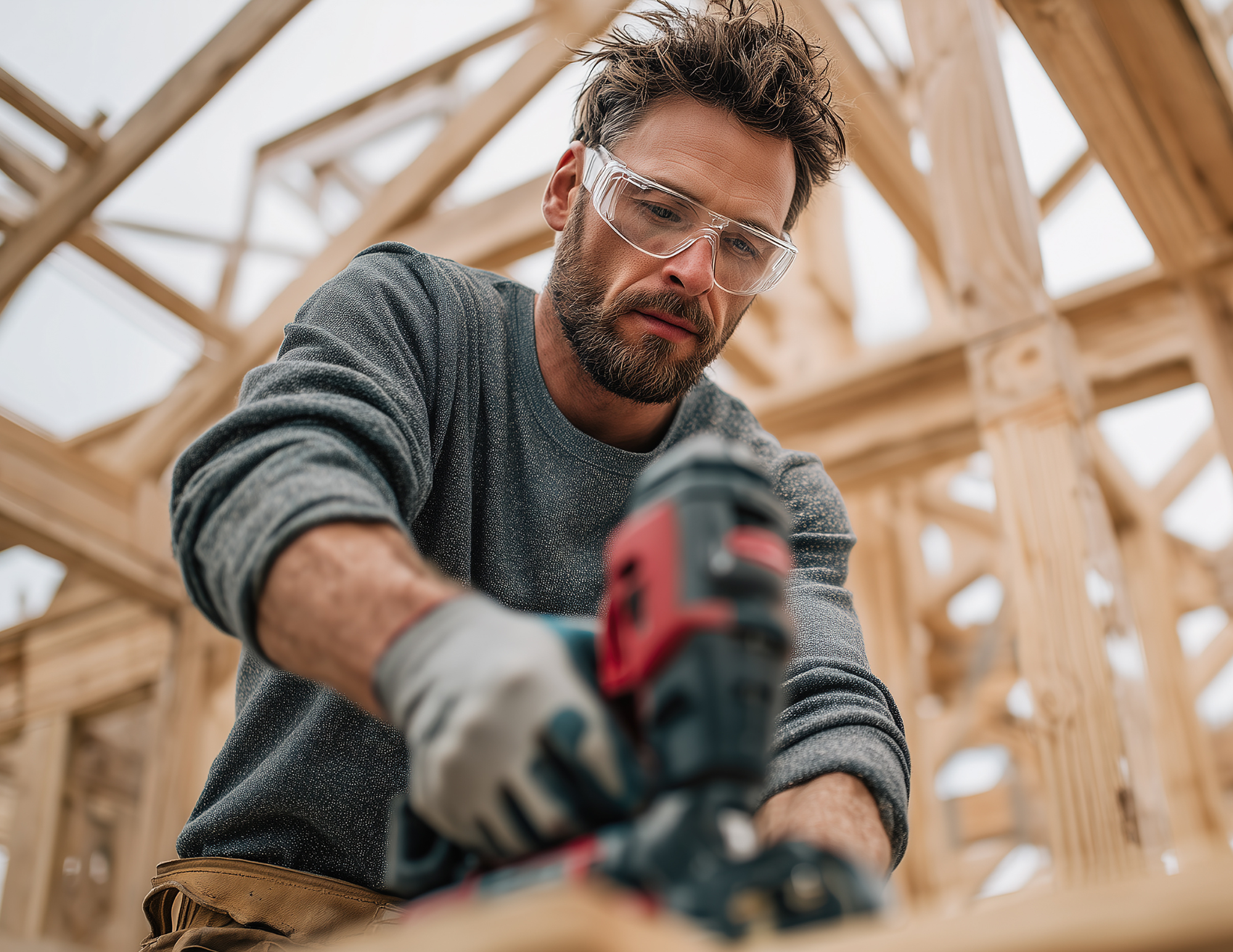 A carpenter uses a power drill against a backdrop of house framing still under construction
