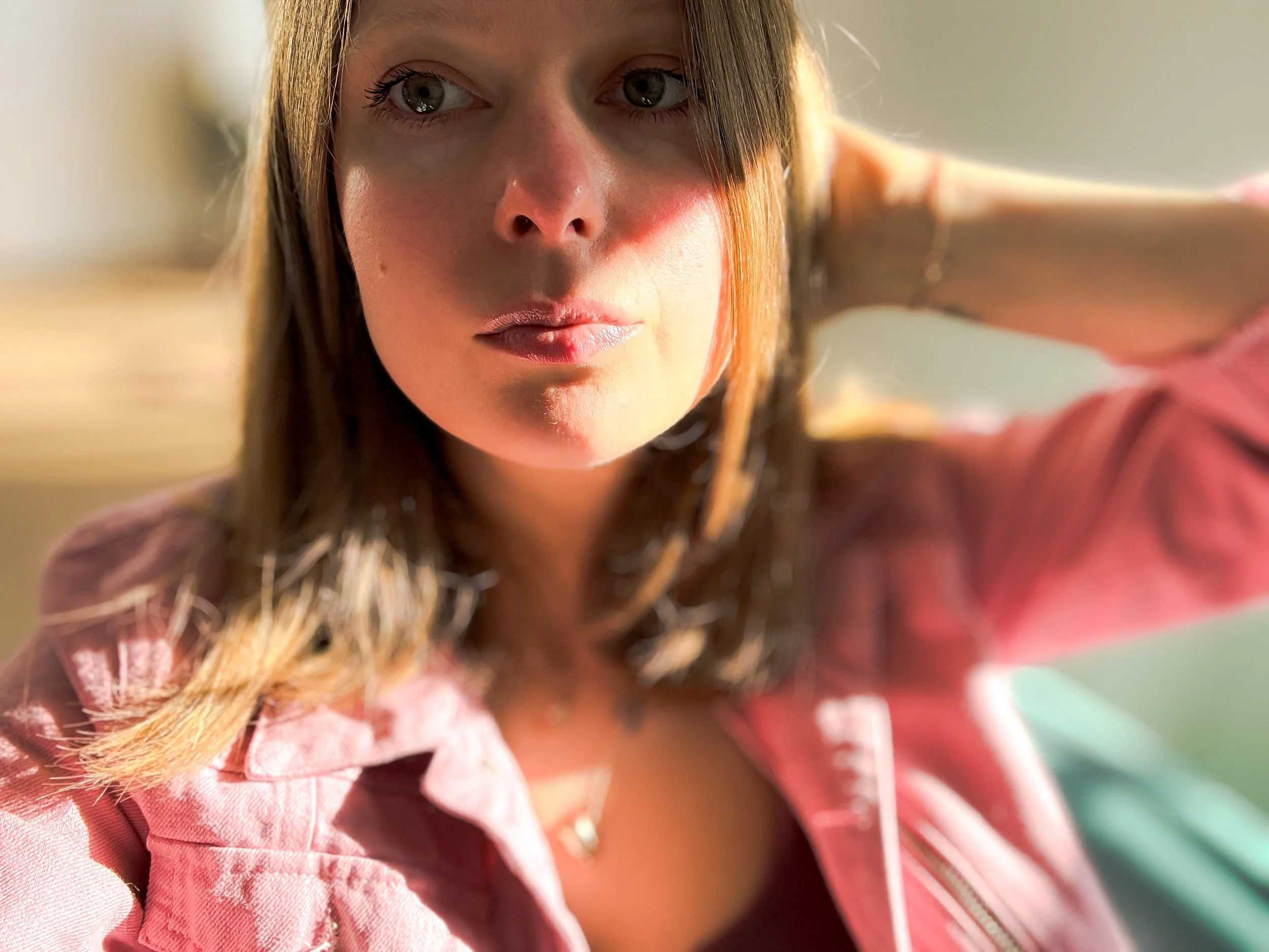 Close-up of a young woman with light brown hair and green eyes, wearing a pink jacket, looking directly at the camera, with sunlight illuminating her face.