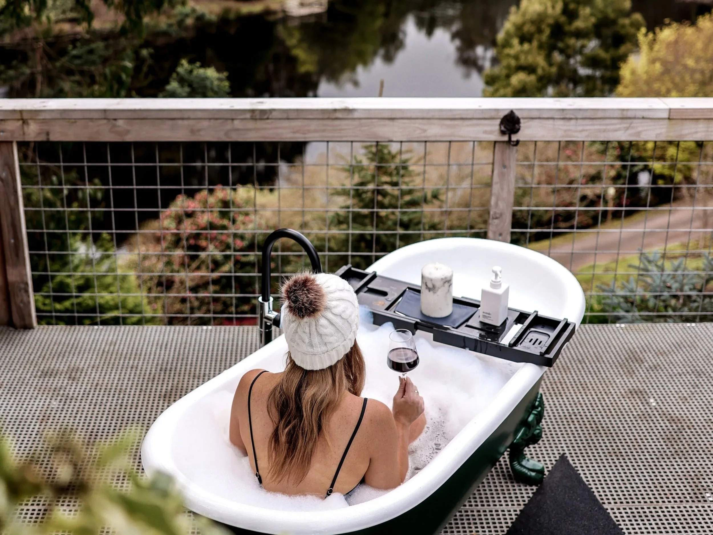 A woman relaxing in a bubble bath on a balcony with a view of a lush, green landscape and lake. She is wearing a white knit hat with a pom-pom, holding a glass of red wine, and facing away from the camera.