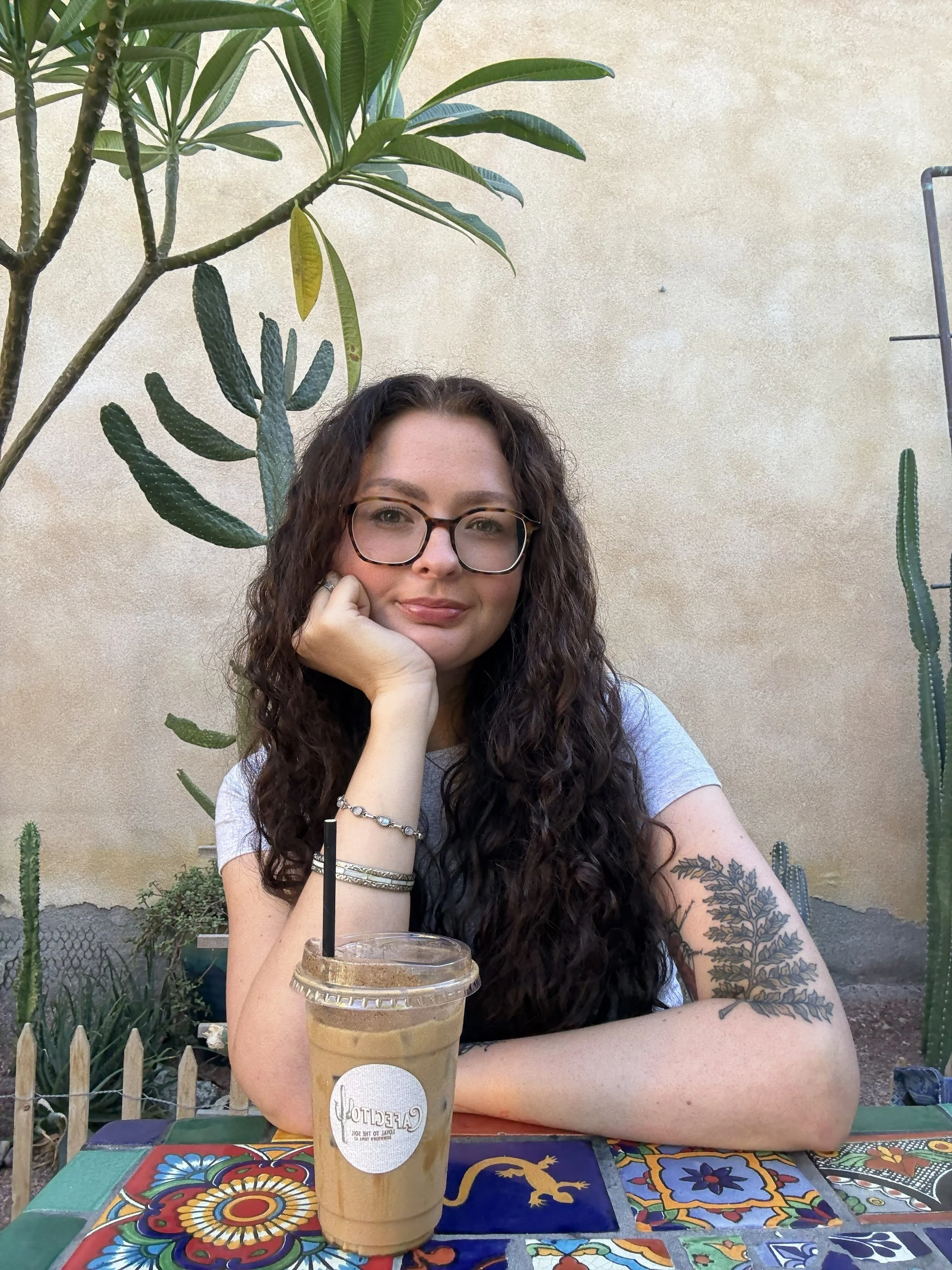 Photo of a young woman with fair skin, long dark curly hair, wearing glasses sitting at a table in front of green plants and a stucco wall