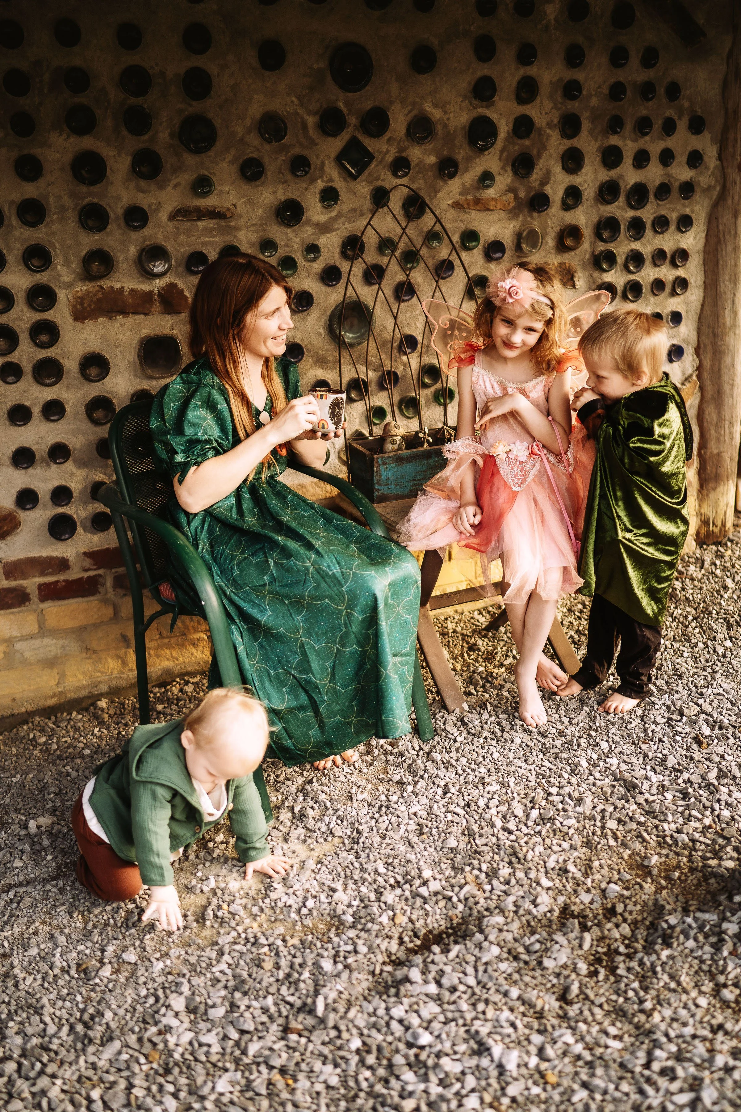 woman in a long green dress holding a mug of coffee, a toddler is playing on the gound in front of her, another child in a pink dress is sitting to her right and a third child in a moss green cape is standing near the child in pink