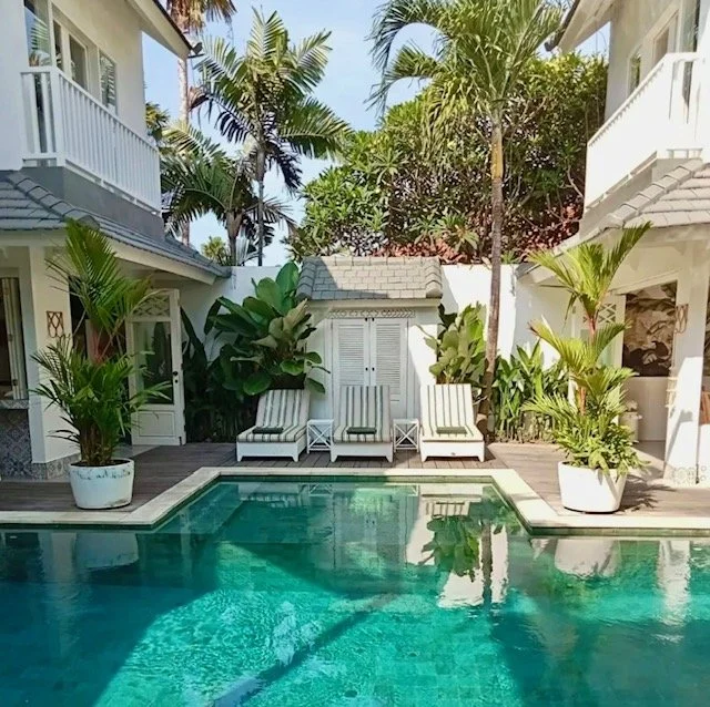 Swimming pool surrounded by white lounge chairs and lush green tropical plants in a luxurious backyard.