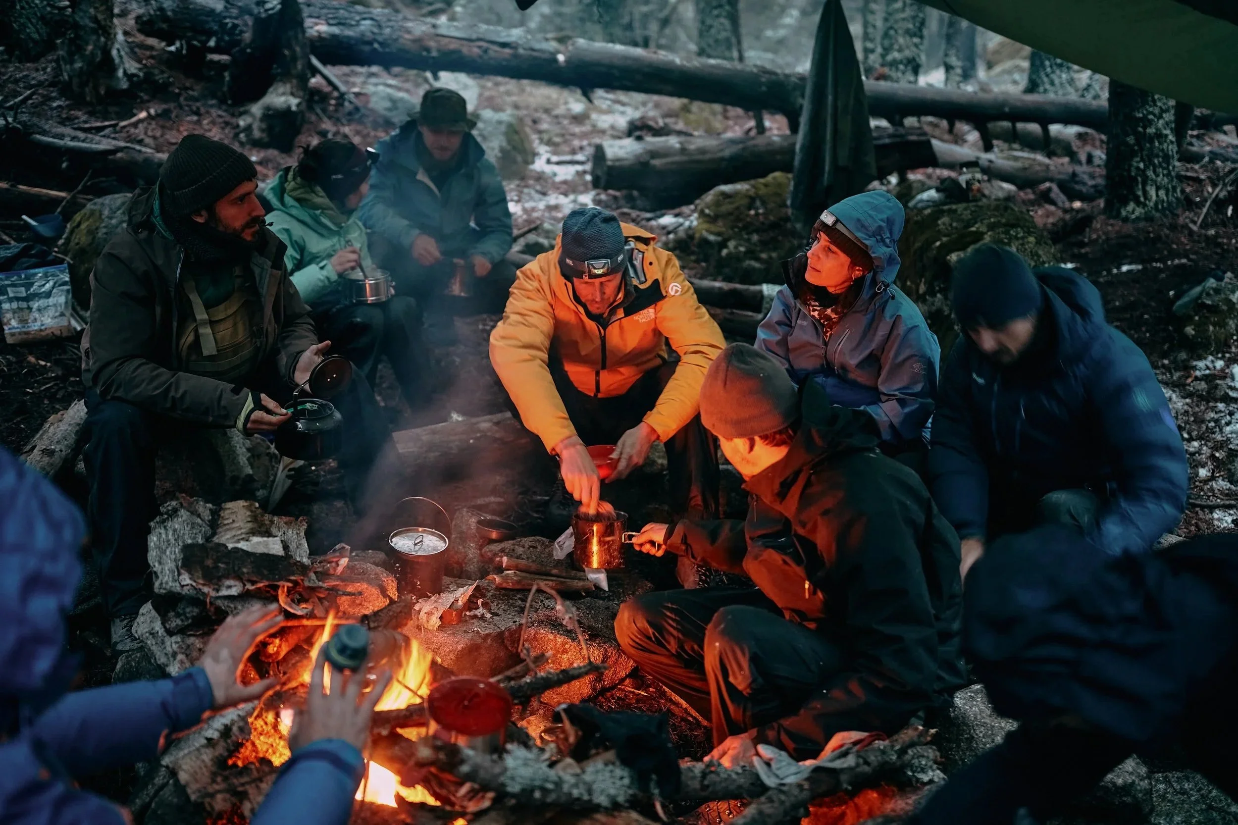 A group of people camping in a forest around a campfire, wearing outdoor gear and headlamps, with trees and logs in the background.