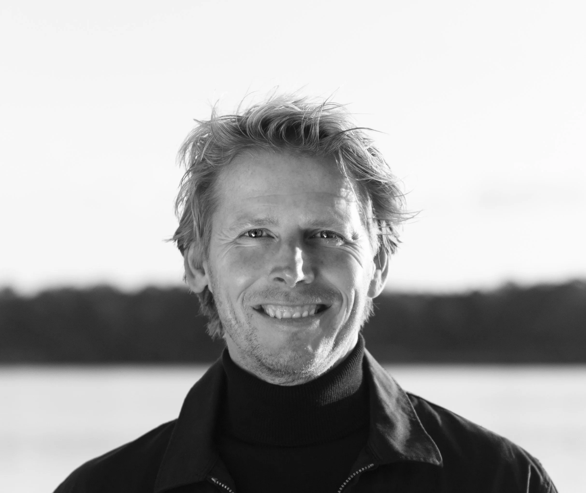 Black and white photo of a smiling man with tousled hair wearing a dark zip-up jacket and turtleneck, standing outdoors against a bright sky