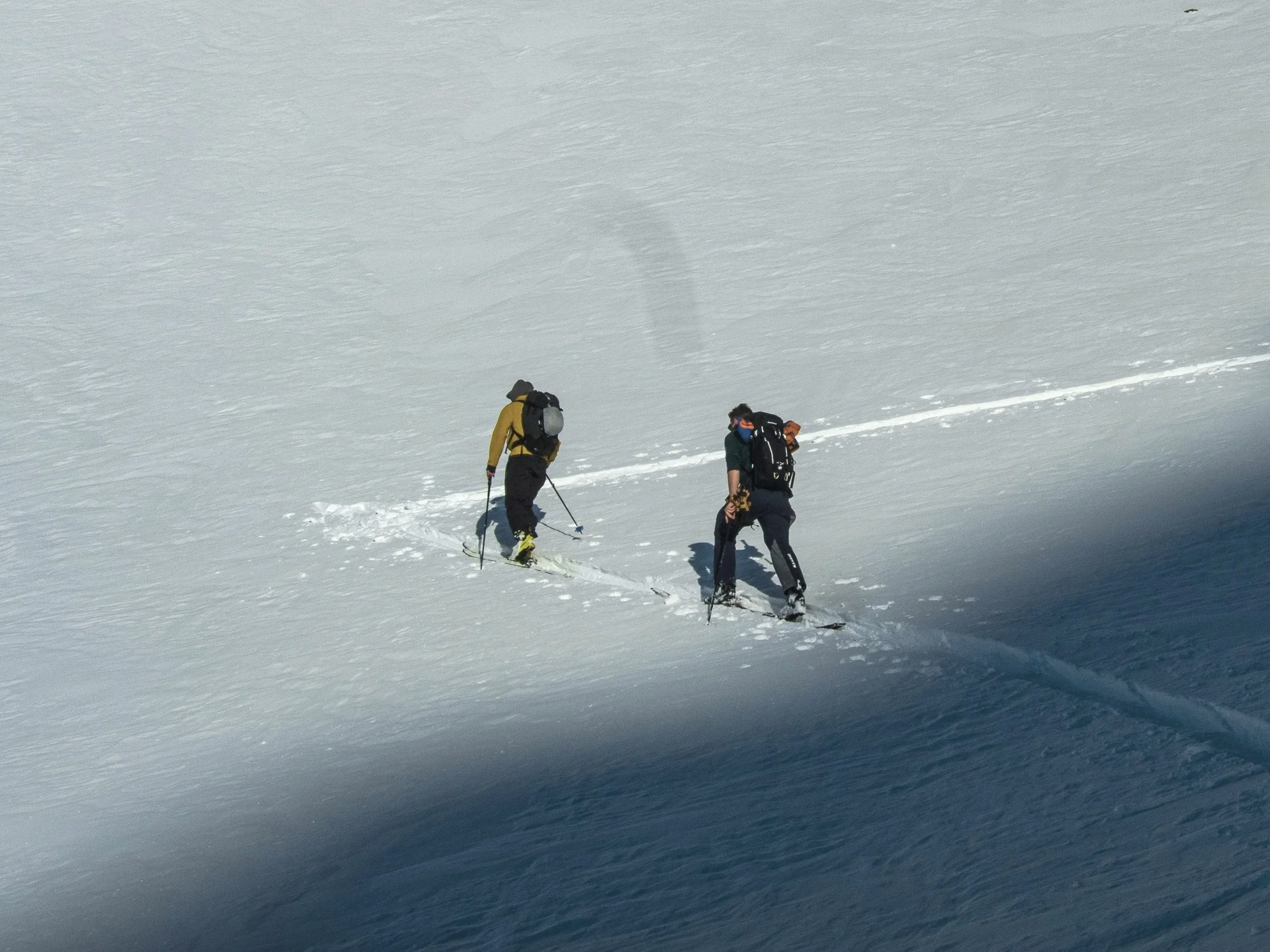 Two people in winter gear, including backpacks, skis, and poles, are walking across a snowy, mountainous landscape under a cloudy sky.