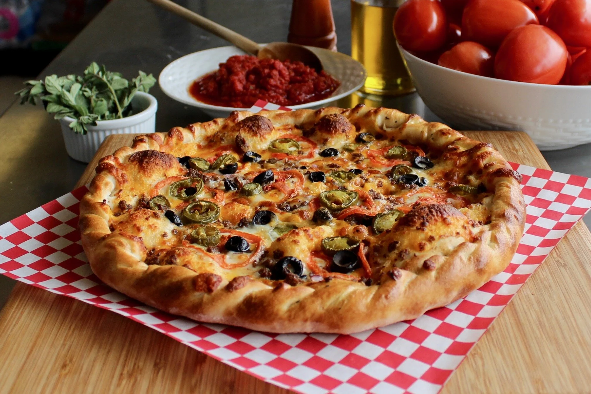 Loaded pepperoni pizza with jalapeños, black olives, peppers, and melted cheese on a checkered paper liner