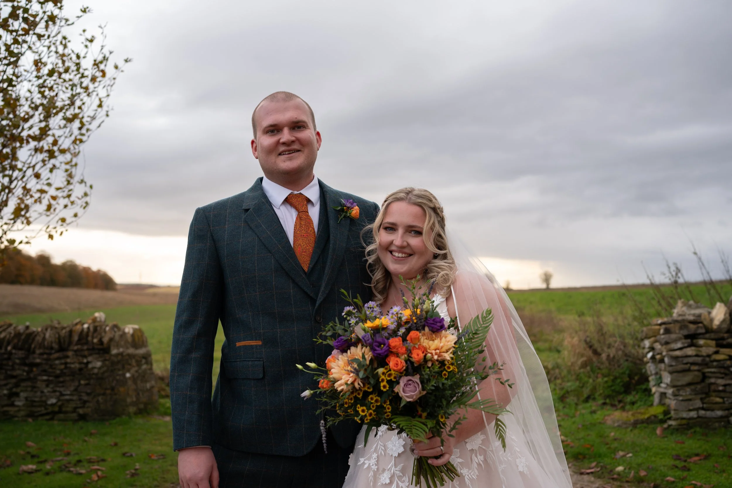 A bride and groom standing outdoors, the groom in a dark suit with an orange tie and the bride in a white dress holding a colorful bouquet. The background features fields and a stone wall under a cloudy sky.