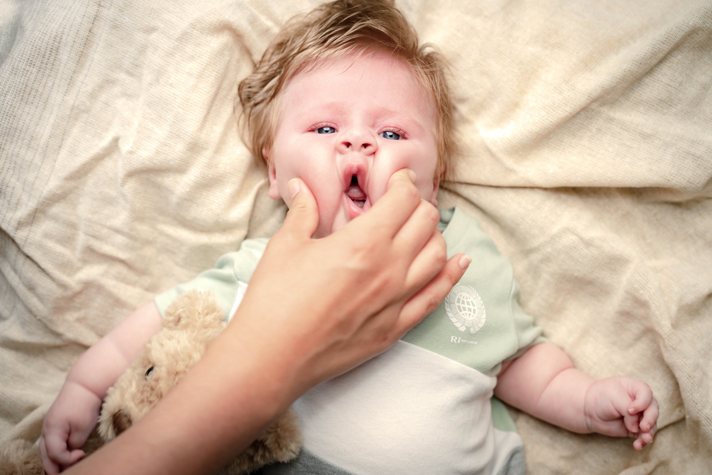 Close-up of a baby lying on a soft beige blanket, with an adult hand gently squeezing the baby's cheeks. The baby has light hair and is wearing a light-colored shirt. A plush toy is partially visible.