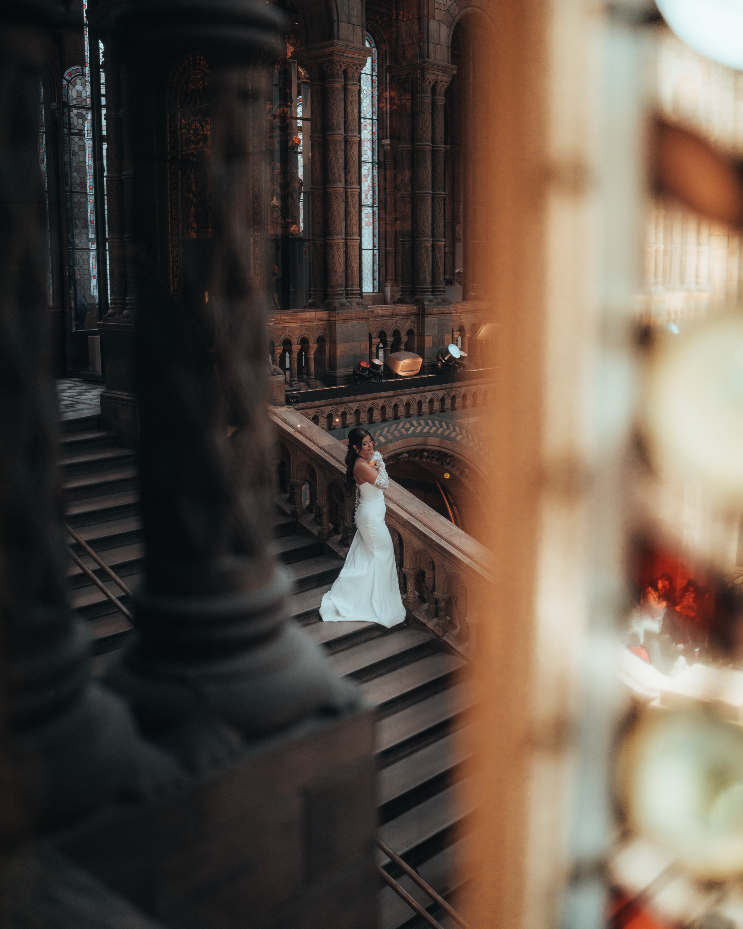 Bride in white dress posing on grand staircase in historic building with arched windows and ornate architecture.